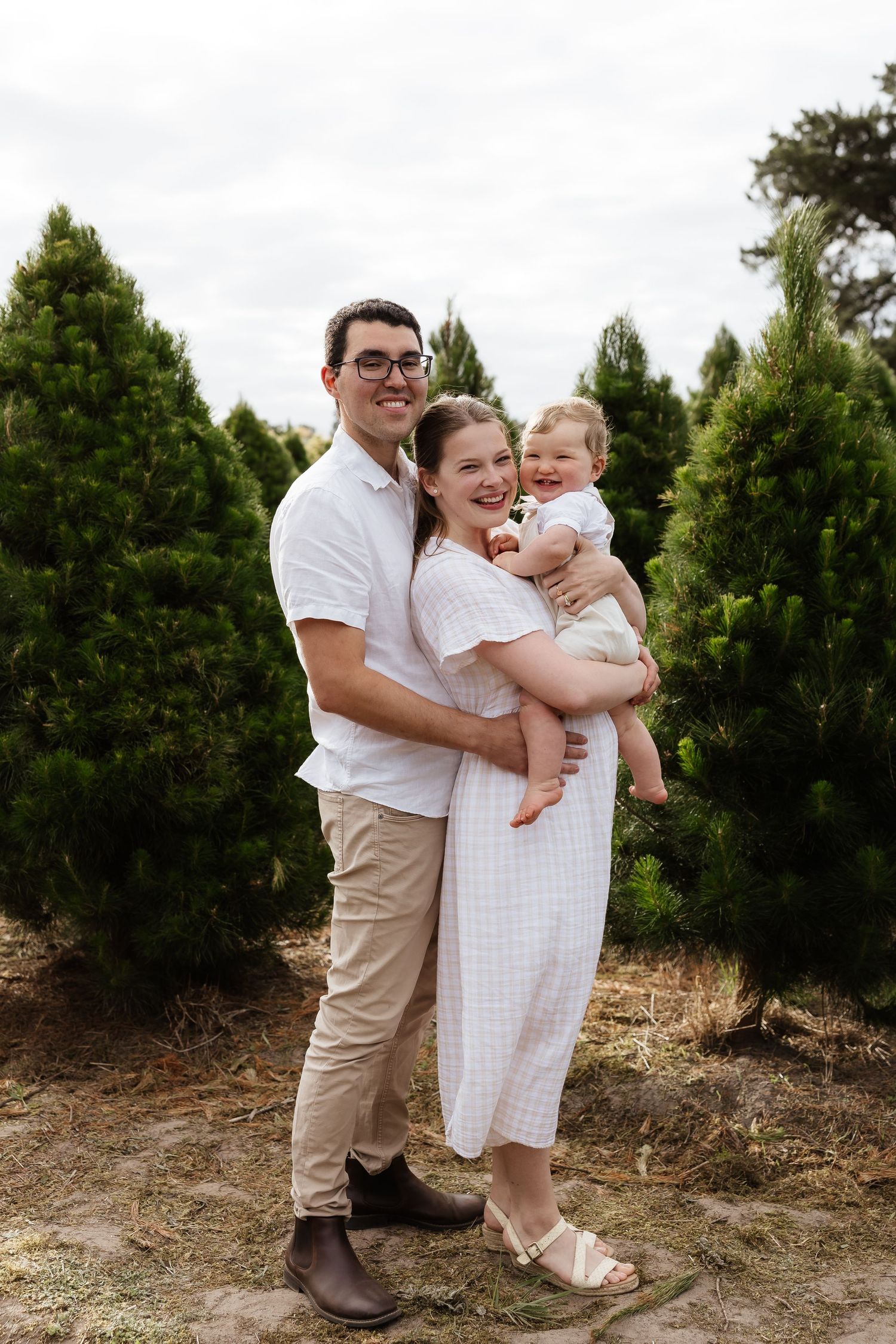 Family in coordinating white and beige outfits pose together outdoors against green trees.