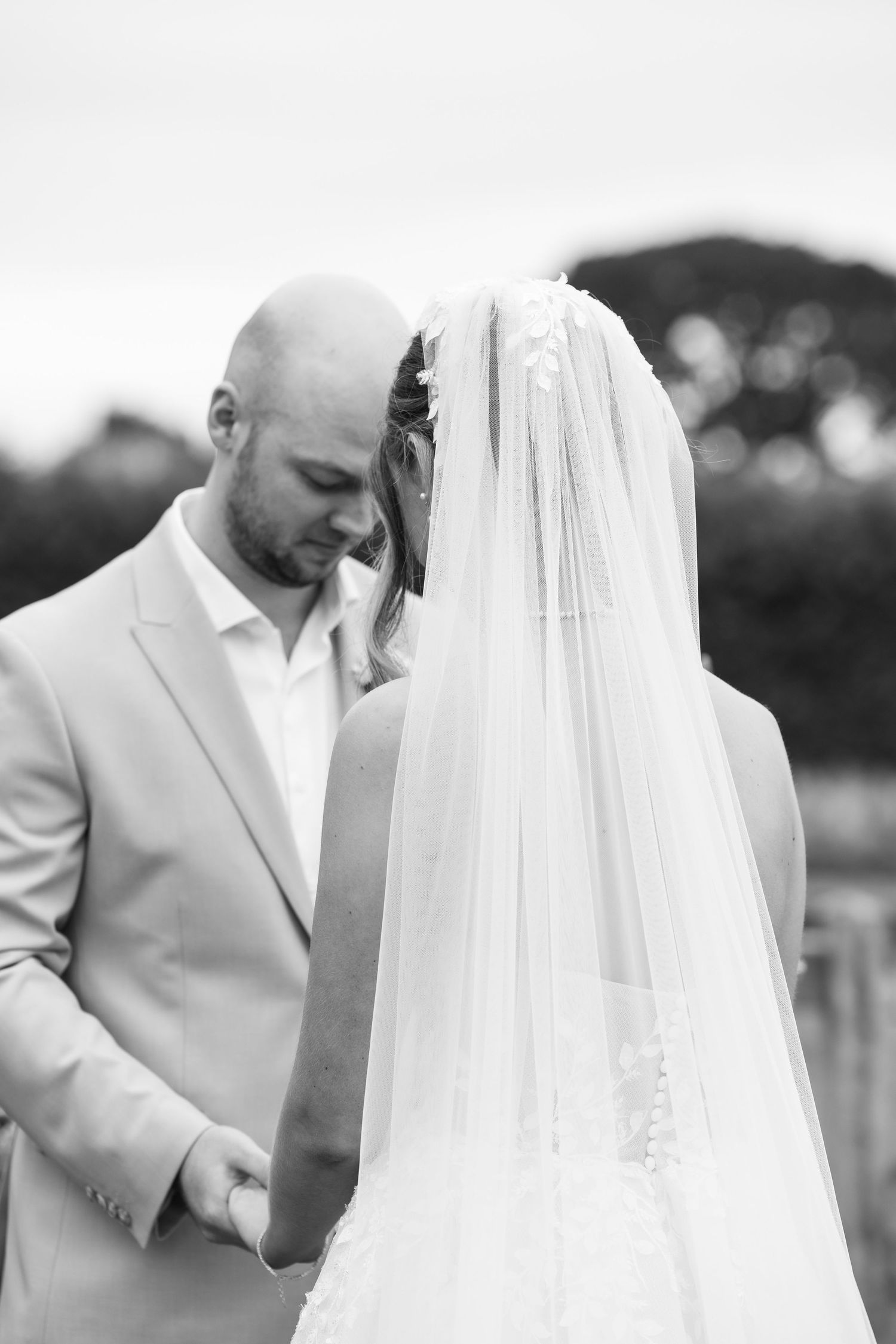 Black and white photo of a groom holding his bride's hands, her veil cascading down her back at their wedding.