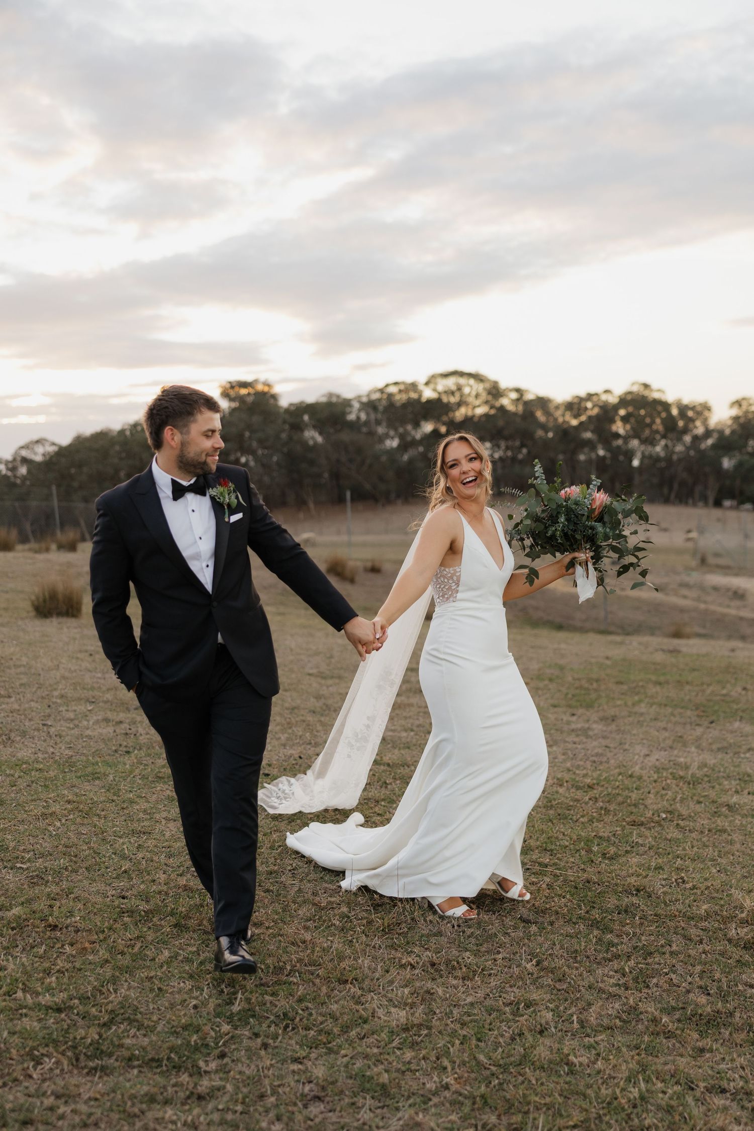 Wedding couple playfully dancing and twirling in an open field at sunset with trees in the background.