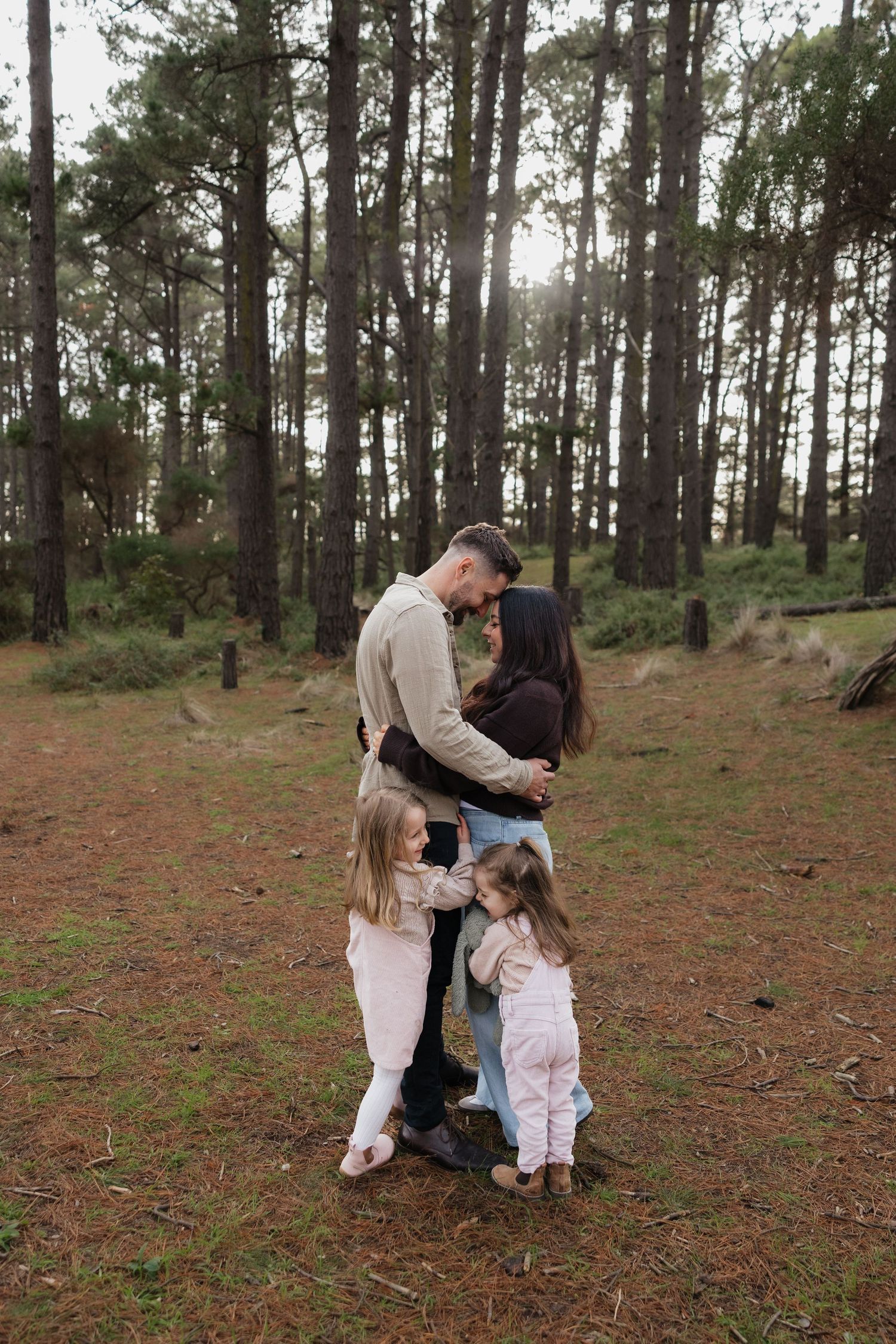 Family of four sharing a tender moment in a sunlit pine forest, with two young girls hugging their parents.