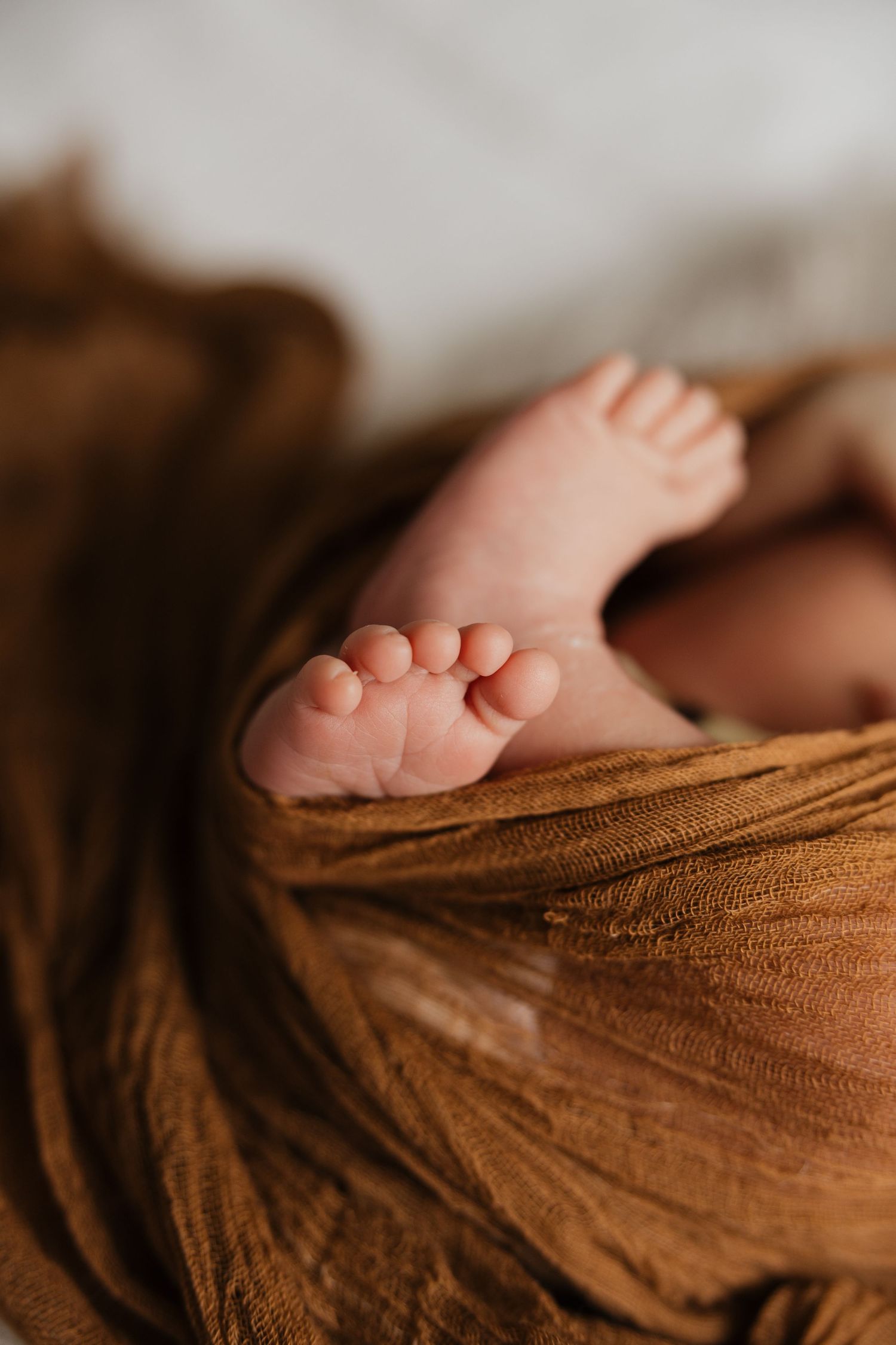 Close-up of tiny newborn baby feet peeking out from a warm rust-colored muslin wrap blanket.