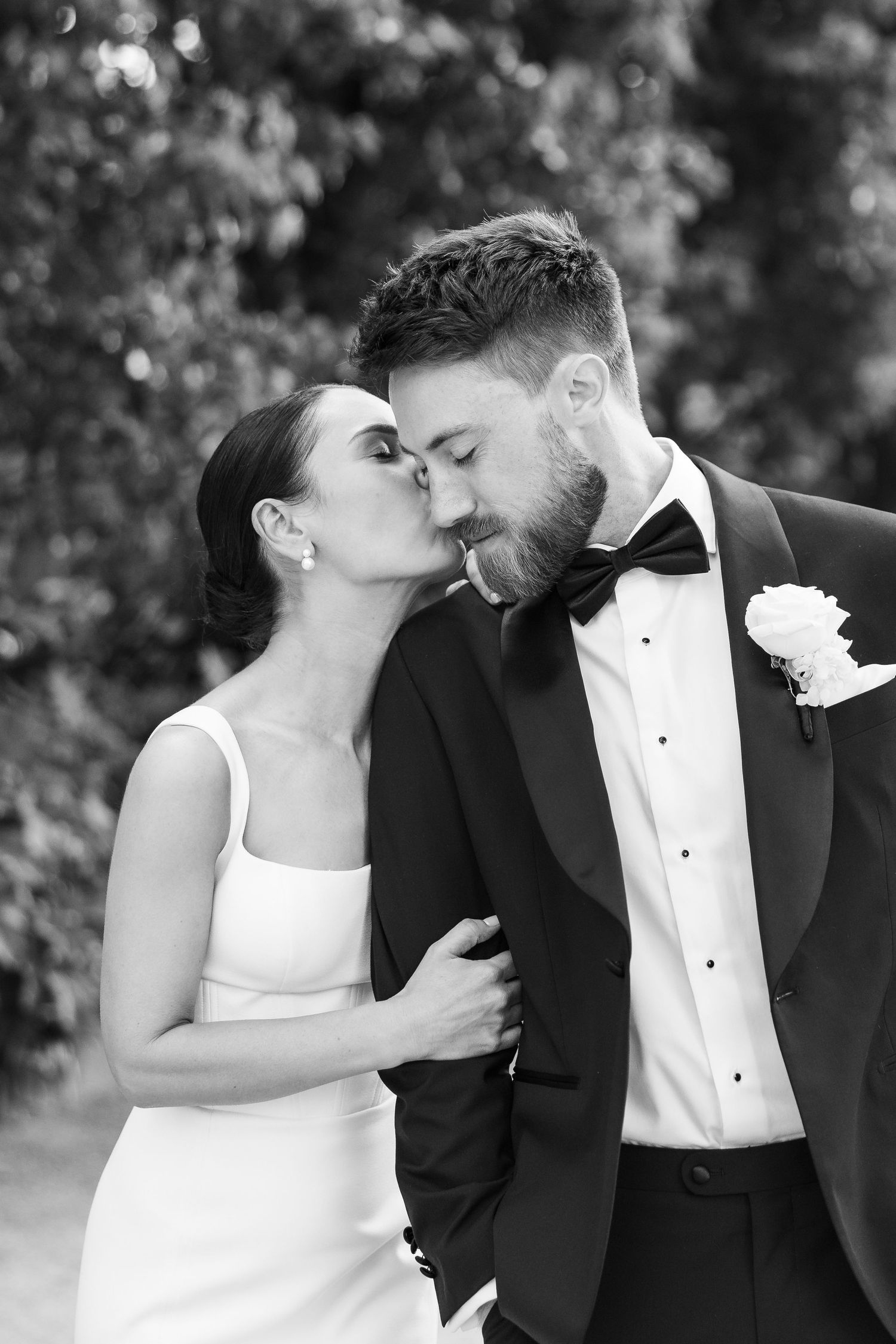 Black and white wedding photo of bride kissing groom in tuxedo outdoors.