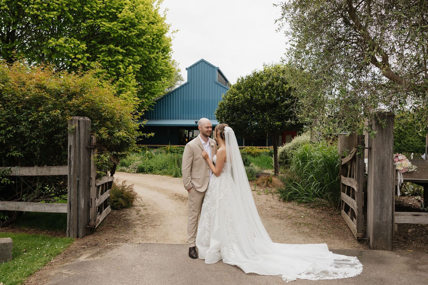 Bride and groom share a romantic kiss on a garden path, with a blue barn and lush greenery in the background.