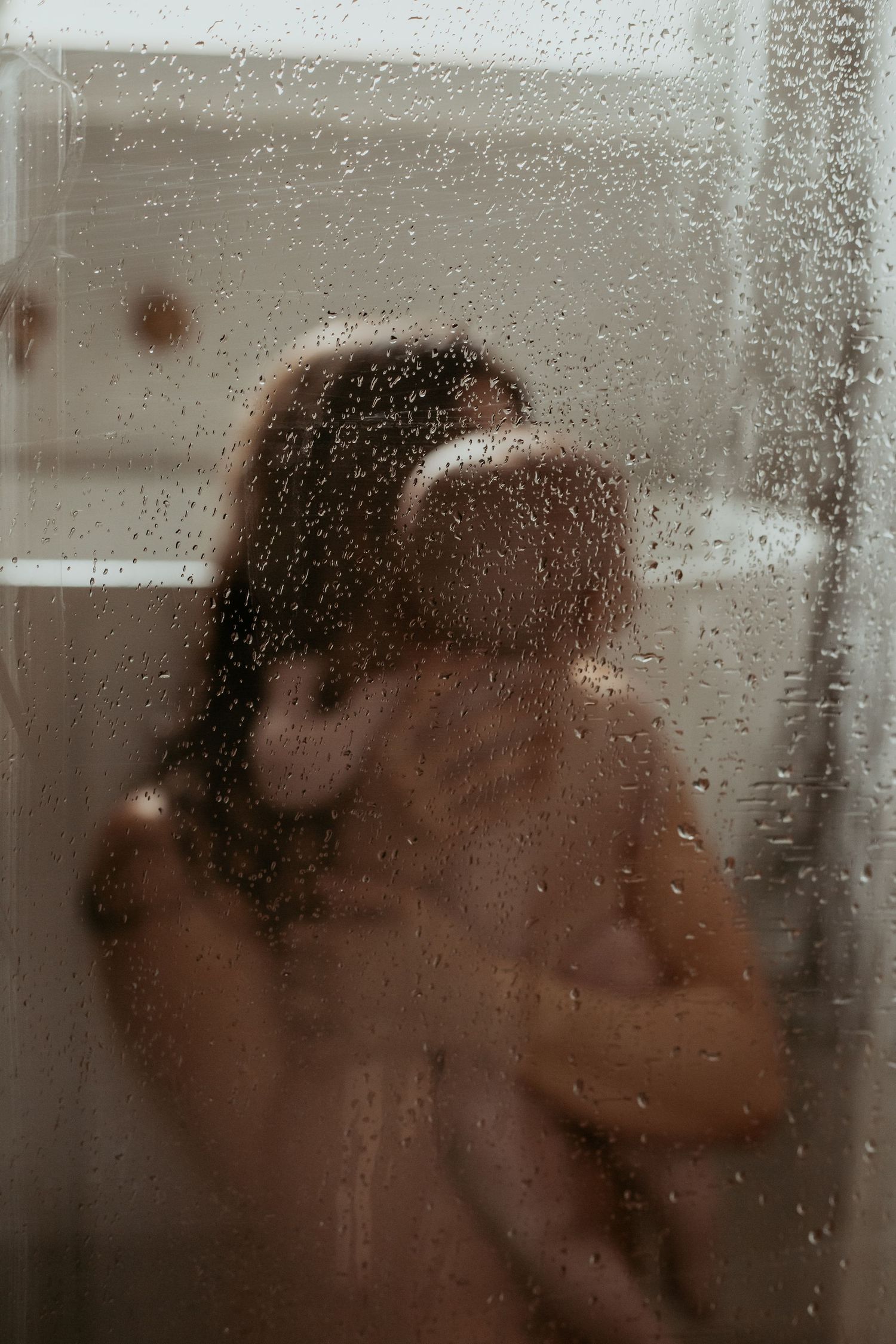 Artistic portrait through rain-covered window with water droplets creating dreamy effect.