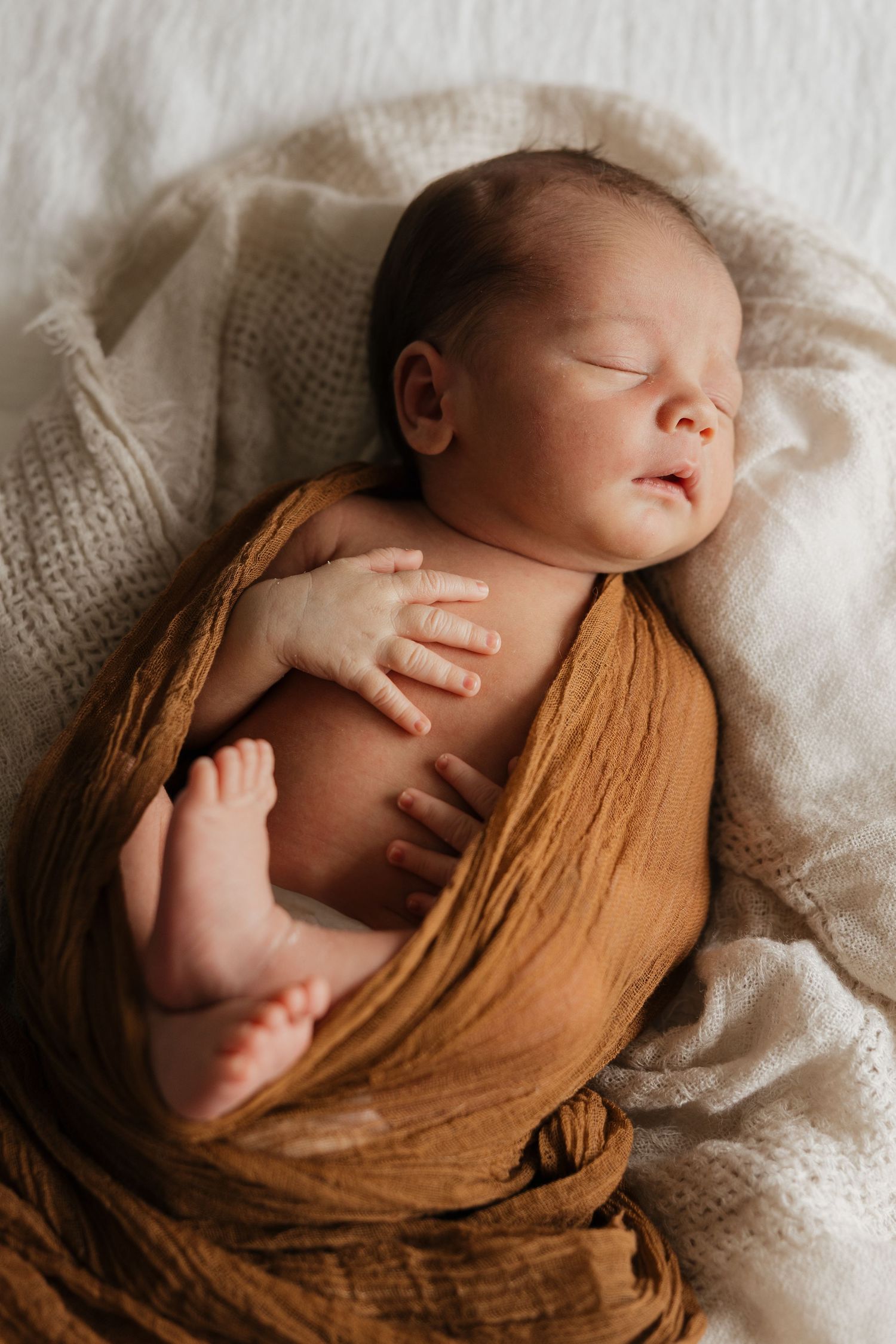 A sleeping newborn swaddled in a soft brown wrap rests peacefully on white textured fabric.
