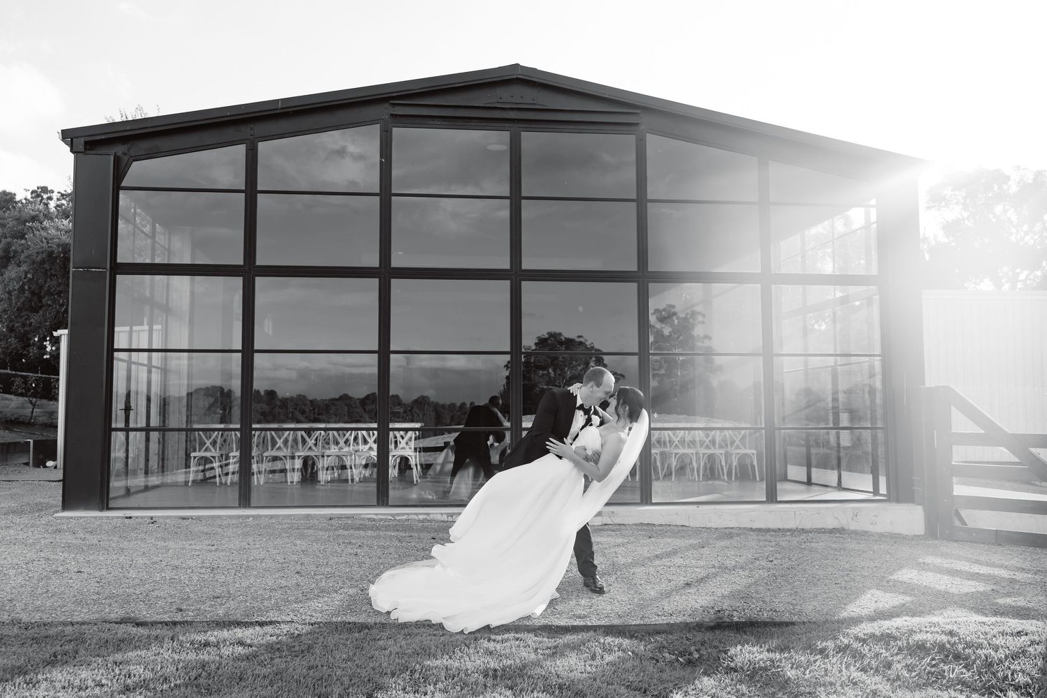 Black and white photo of a bride and groom sharing a romantic dip kiss outside a glass pavilion wedding venue.