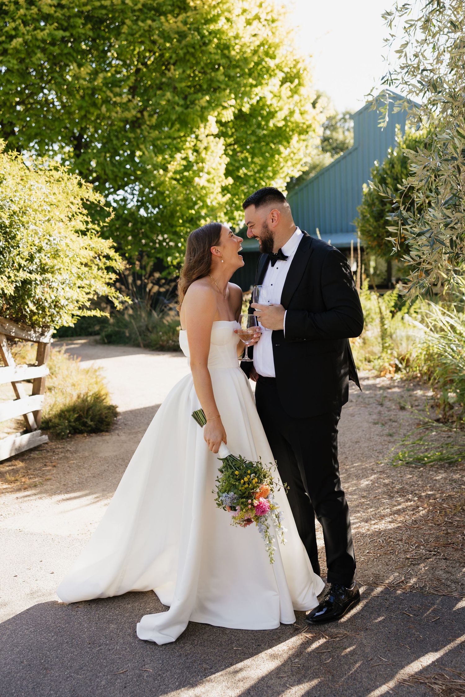Bride and groom share a romantic moment outdoors, she holds a colorful bouquet in a strapless gown, he wears a black tuxedo.