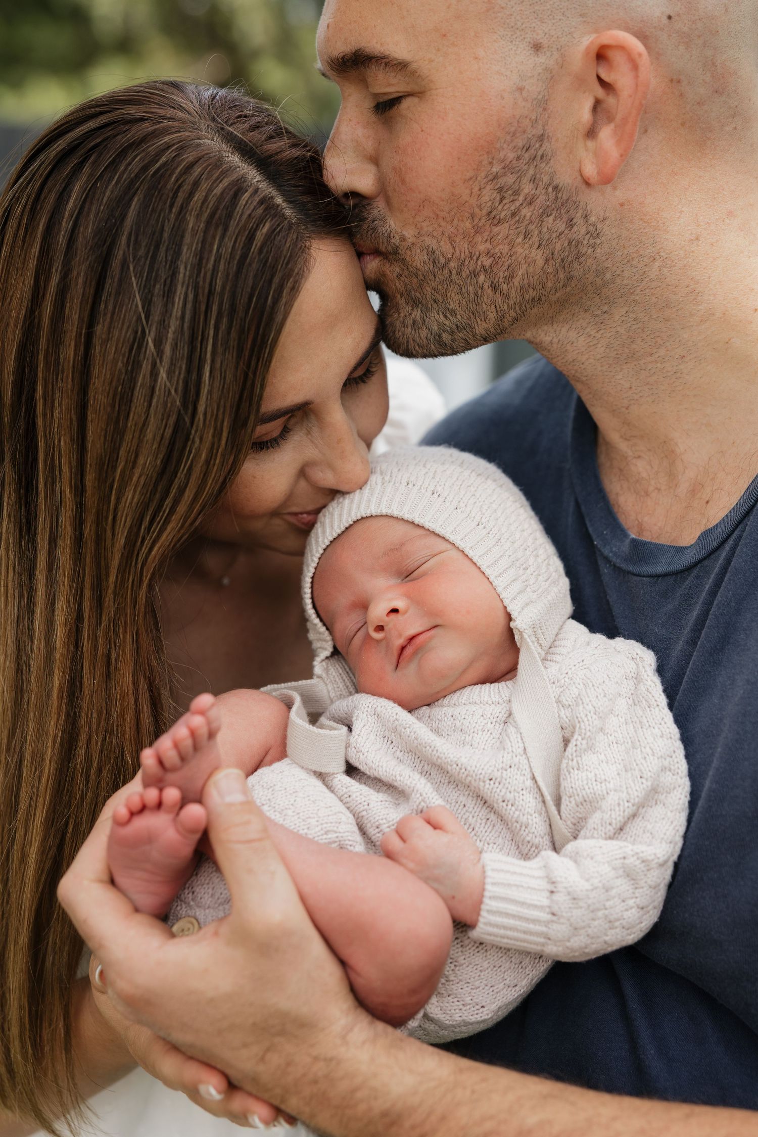 Parents tenderly hold their sleeping newborn baby, with father kissing mother's head in a loving family portrait.