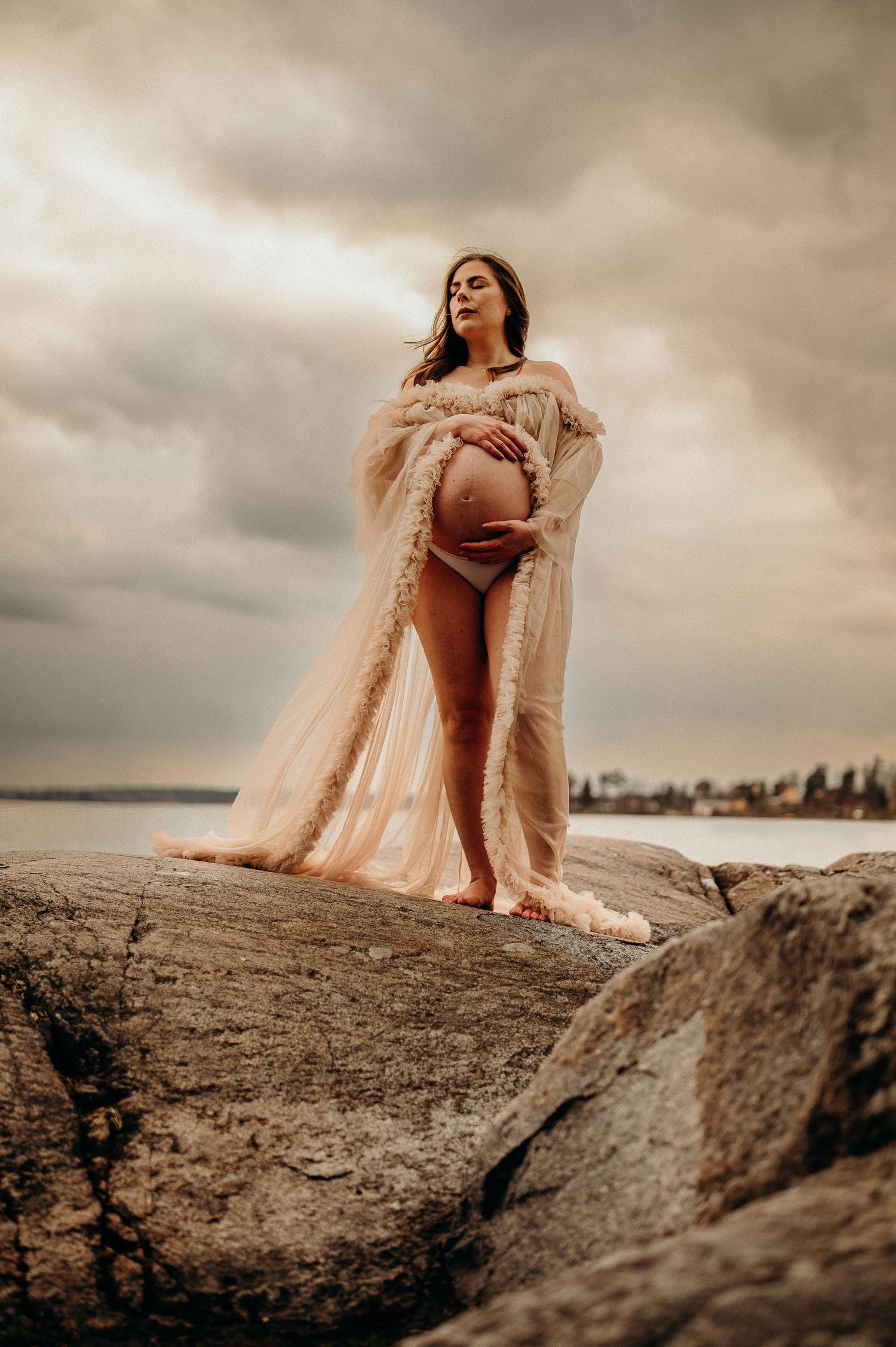 Gravid kvinna i tyllklänning står på en klippa, blundar och håller om sin mage medan dramatiska moln drar förbi – gravidfotografering Sthlm