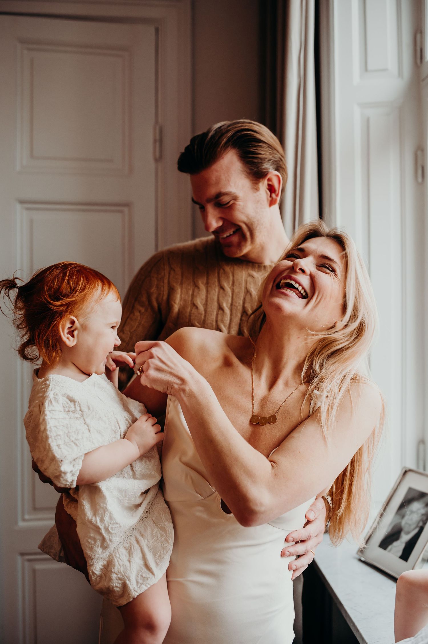 Avslappnad och rolig familjefotografering i hemmet. Mamma, pappa och dotter håller om varandra och skrattar tillsammans.