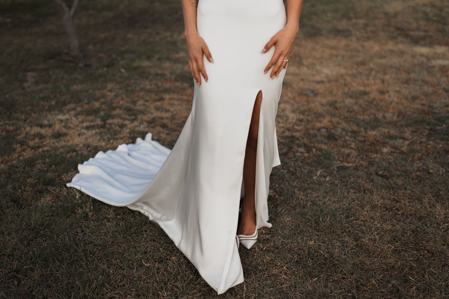 Close-up of elegant white wedding dress with high slit on outdoor field showing flowing fabric detail.