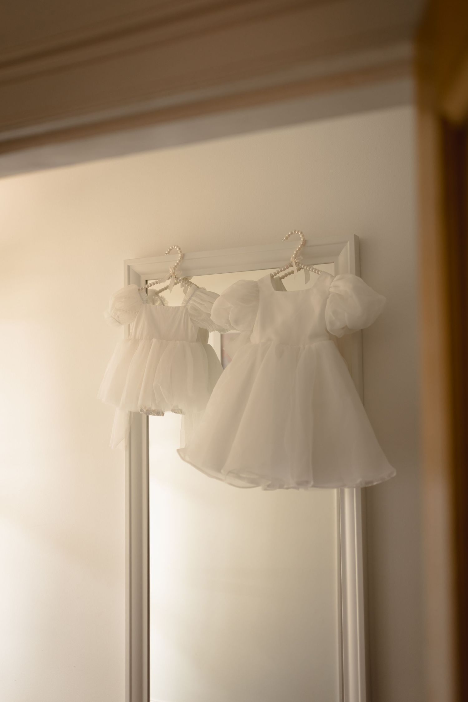 Two delicate white tulle baby dresses hanging on hangers against a light-colored wall in soft, dreamy lighting.
