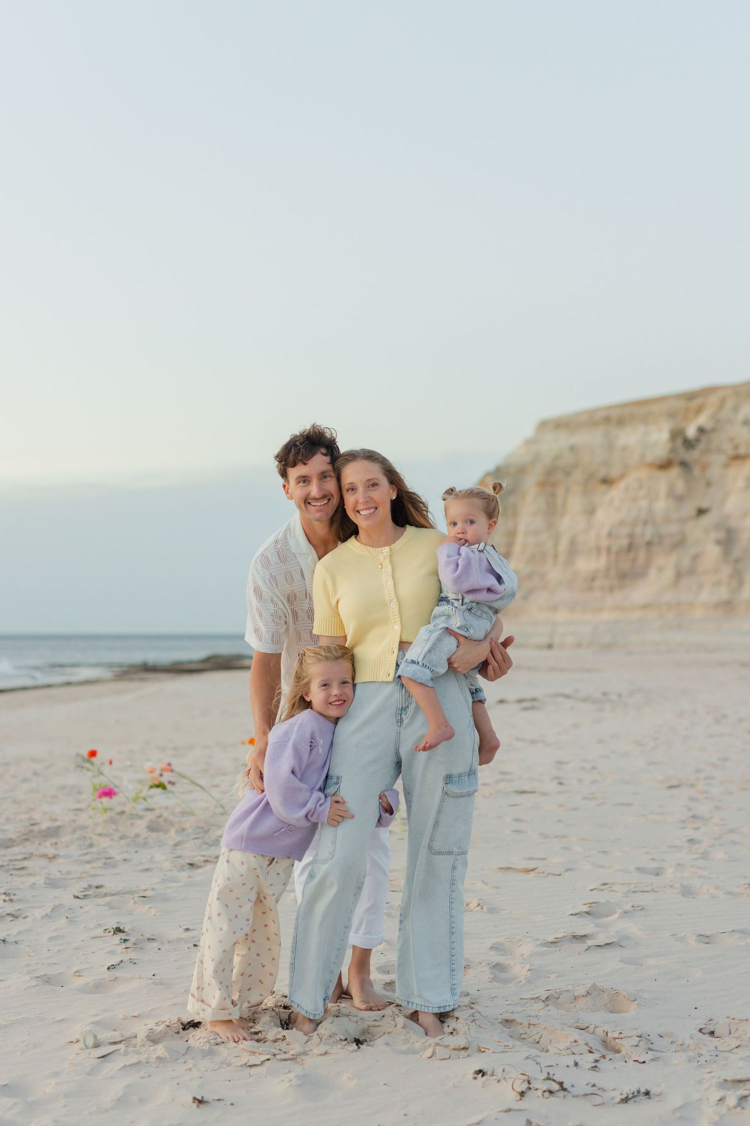 Happy family of four posing on a sandy beach at sunset, parents holding young children near coastal cliffs.