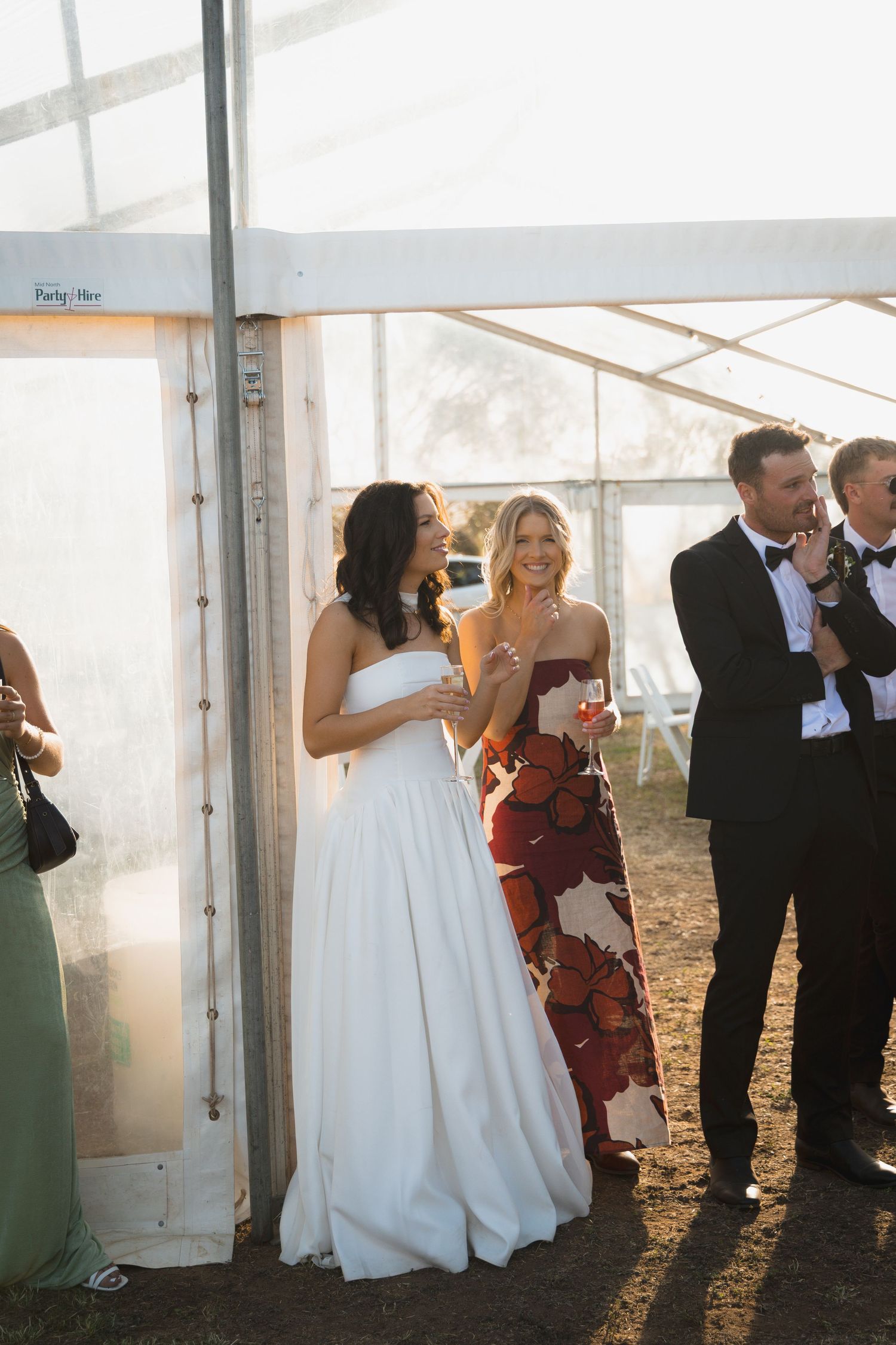 Bride in white gown laughing with guests in floral dress and tuxedo inside a sunlit wedding tent.