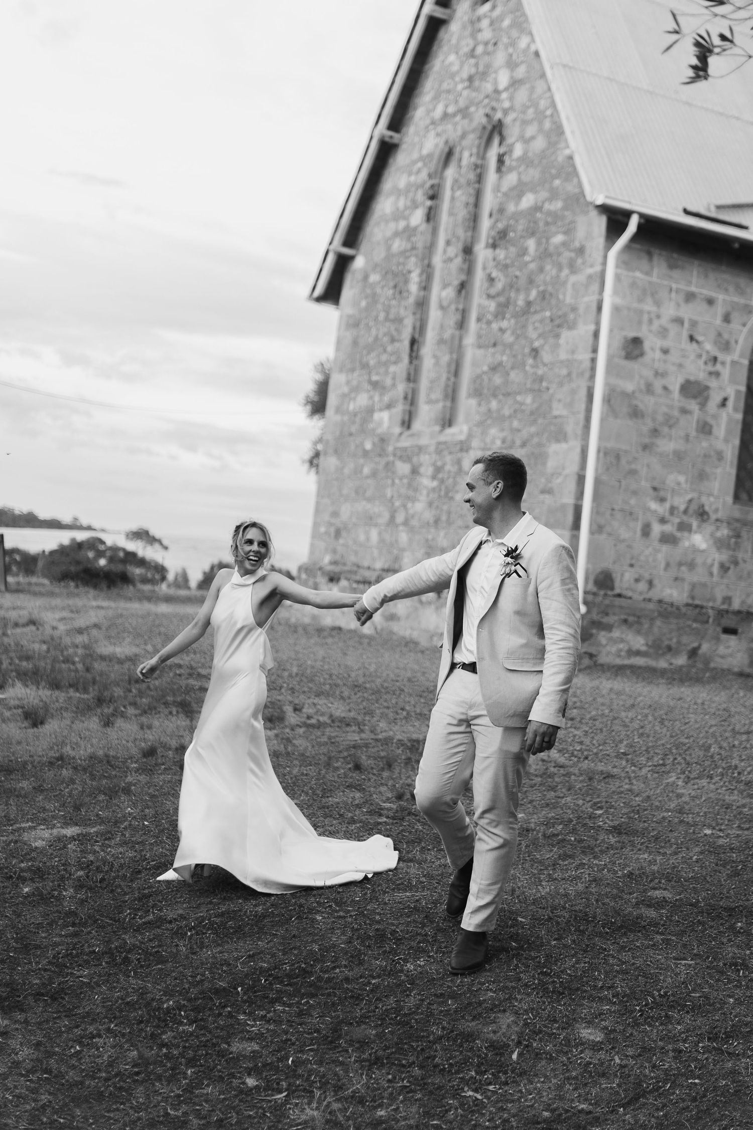 A couple in wedding attire dance and hold hands outside a stone church building in black and white photography.