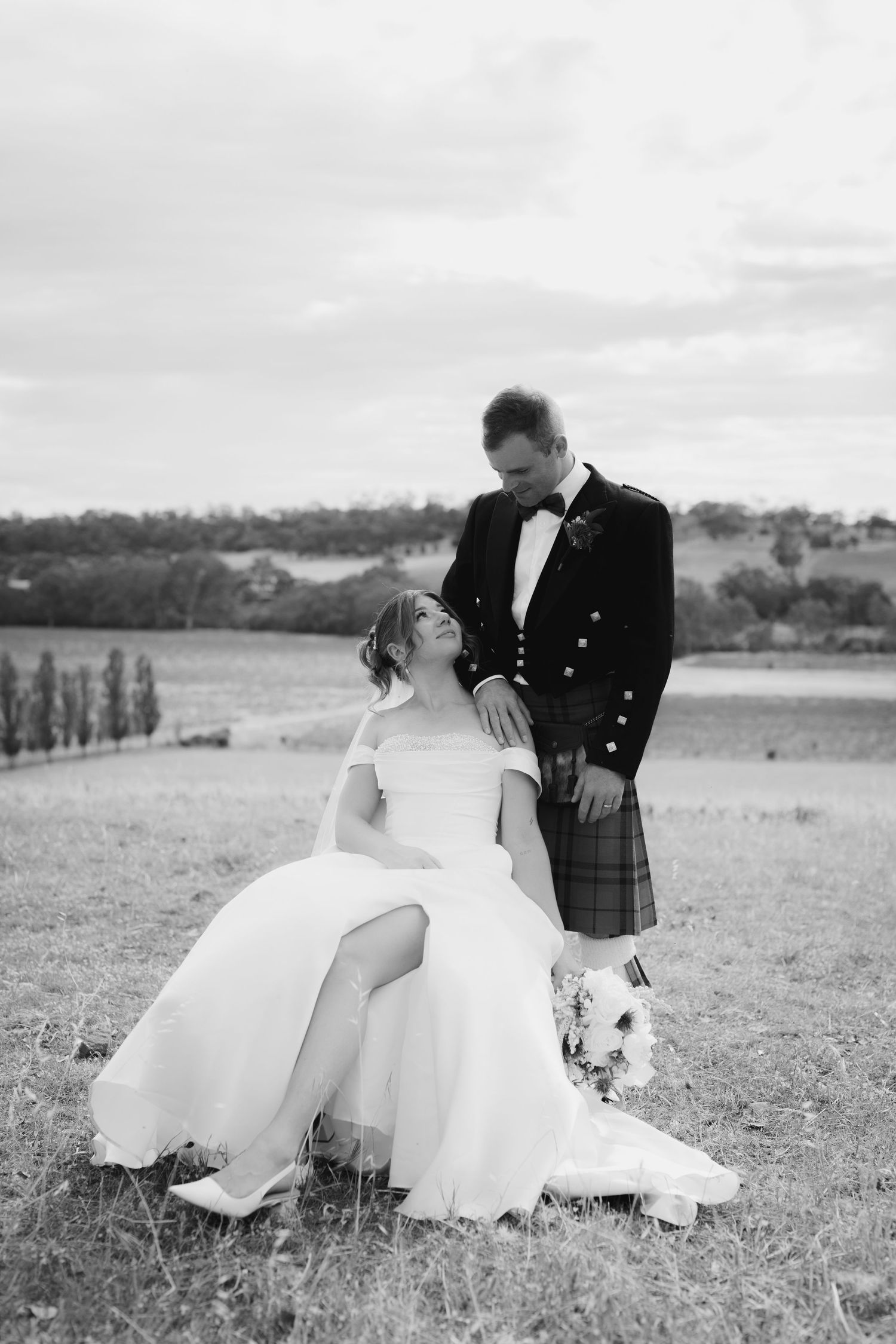 Black and white photo of bride sitting in field as groom in tartan kilt stands beside her holding bouquet.