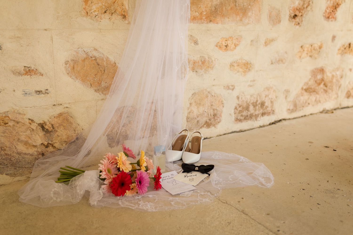A white wedding veil drapes over bridal shoes and a colorful floral bouquet against a textured stone wall.