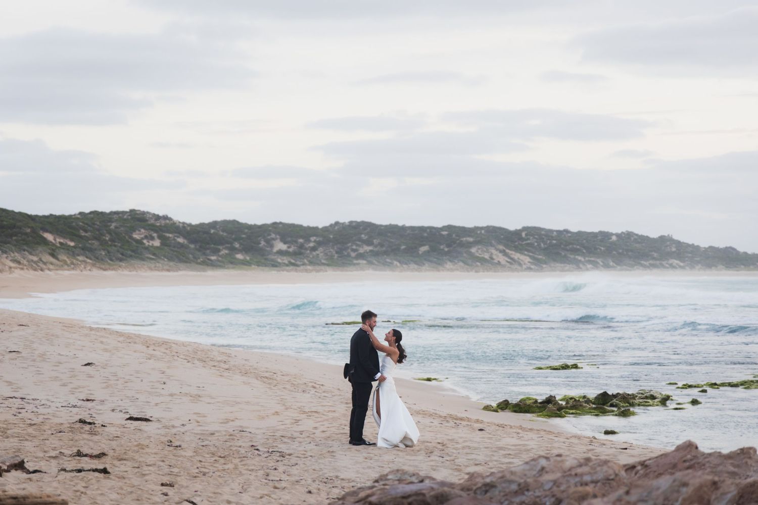 Two people in formal attire stand together on a misty beach with rolling waves and cliffs in the background.