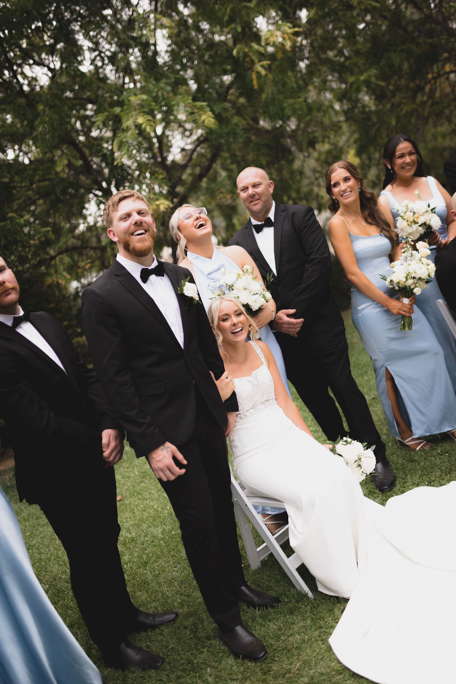 Wedding party gathered outdoors sharing a candid laughing moment during formal photos in elegant attire.