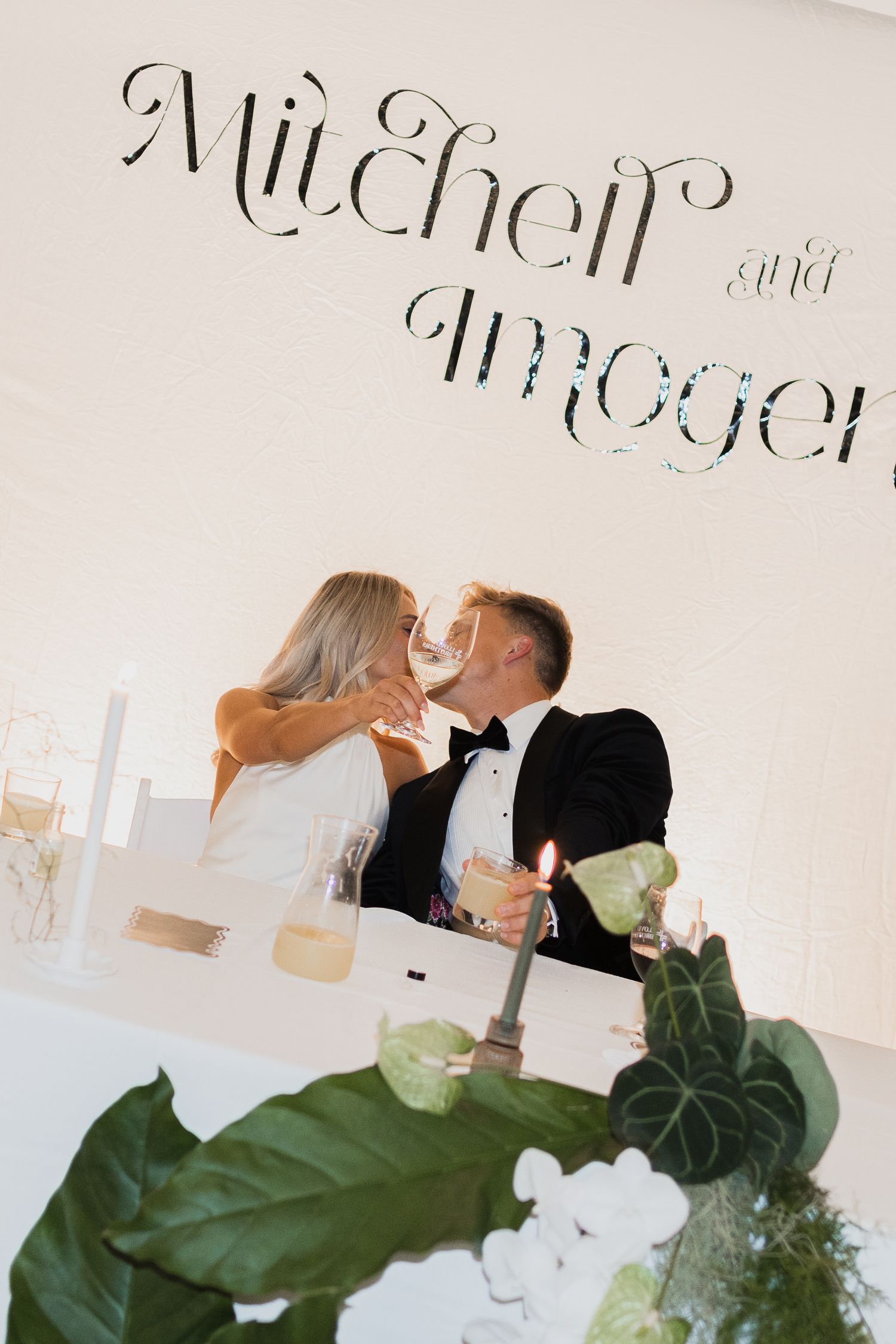 Wedding reception scene with romantic couple sharing intimate moment near decorative greenery and signage.