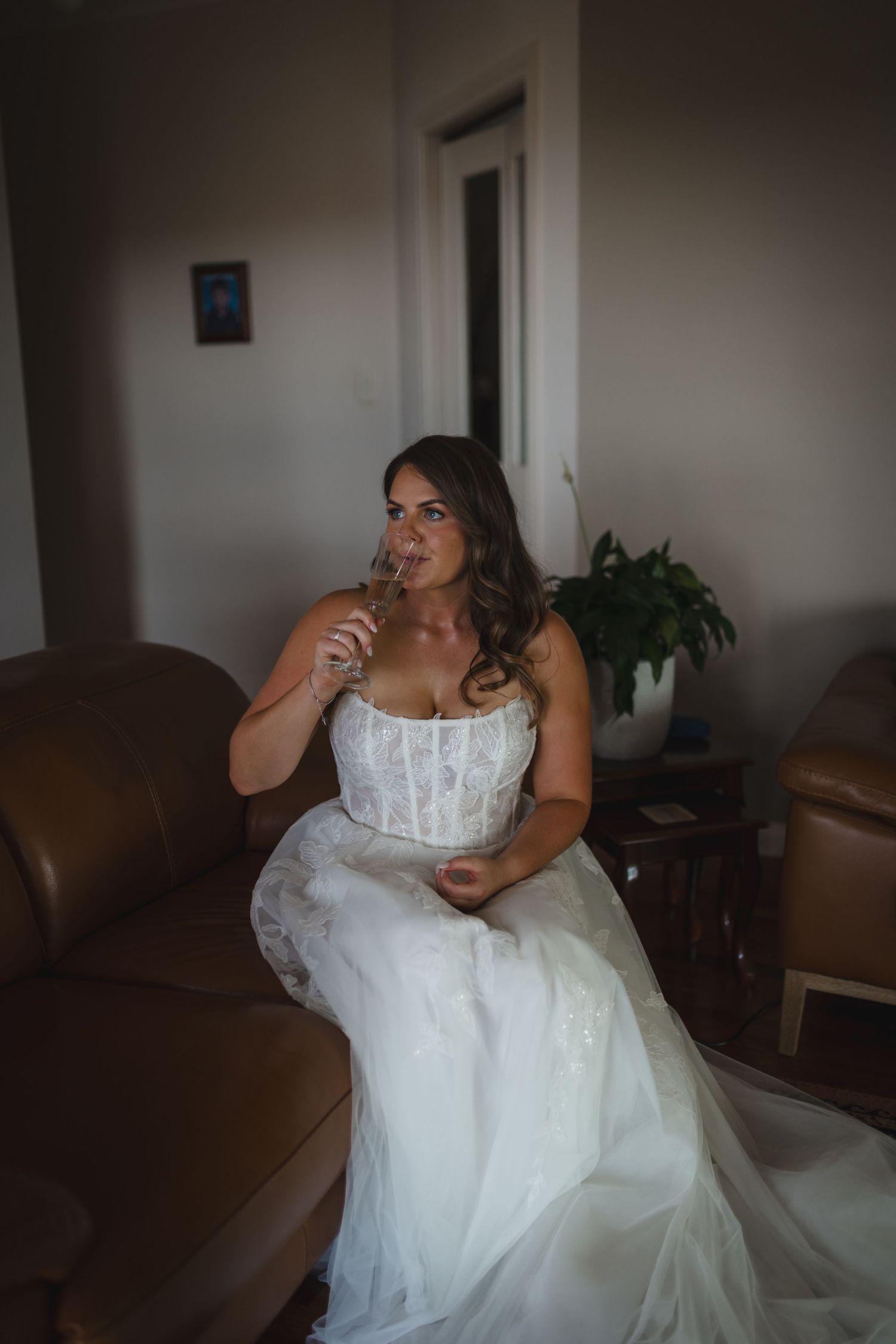A person in a white lace wedding dress sits thoughtfully on a brown leather couch in a dimly lit room.