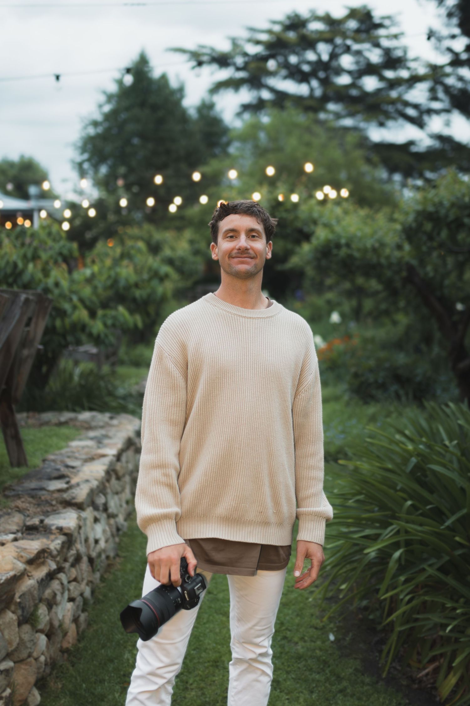 Smiling man in beige sweater holding a camera stands in a lush garden with string lights at dusk.