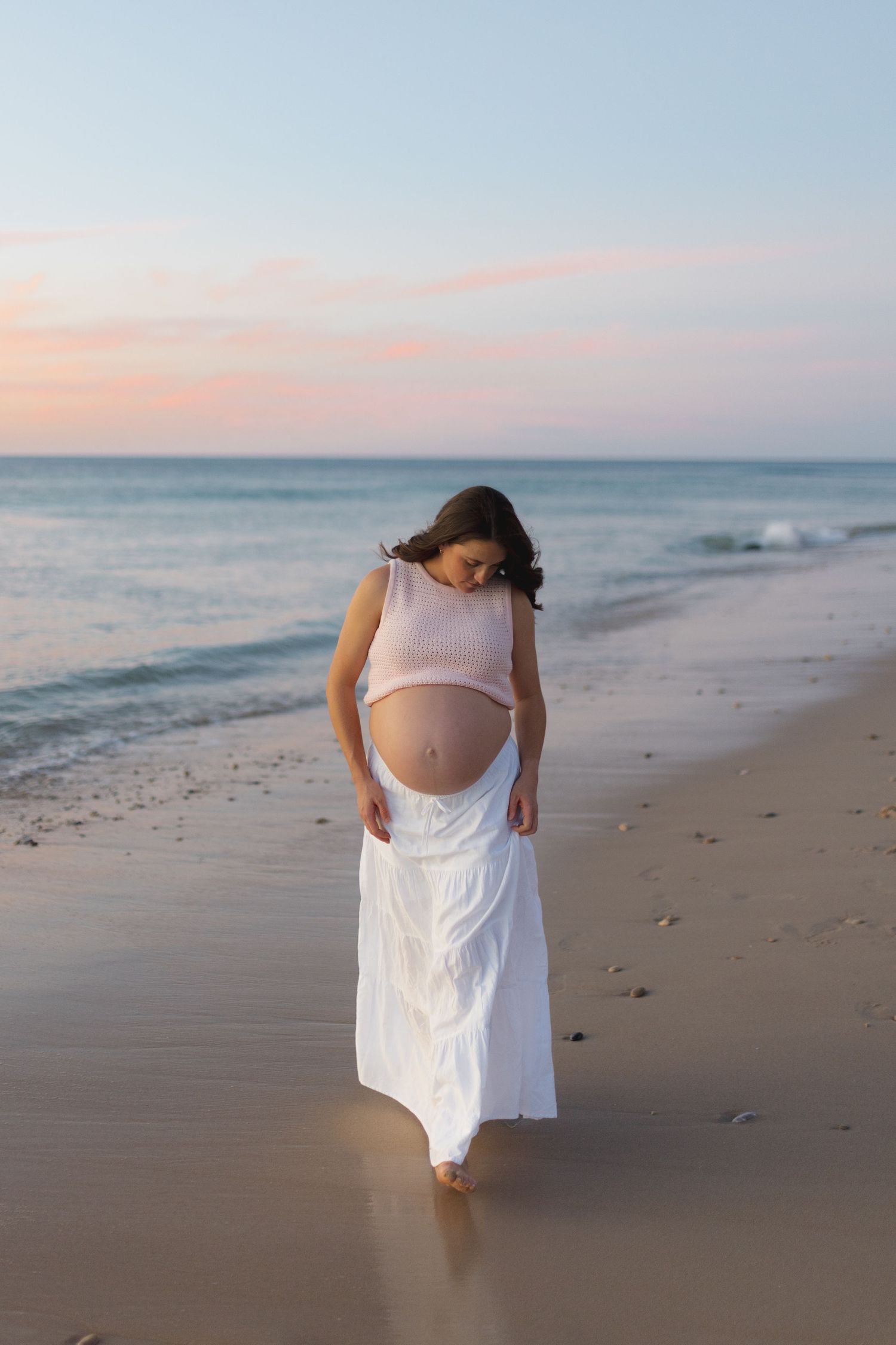 Pregnant woman in a white dress walking along a beach at sunset.
