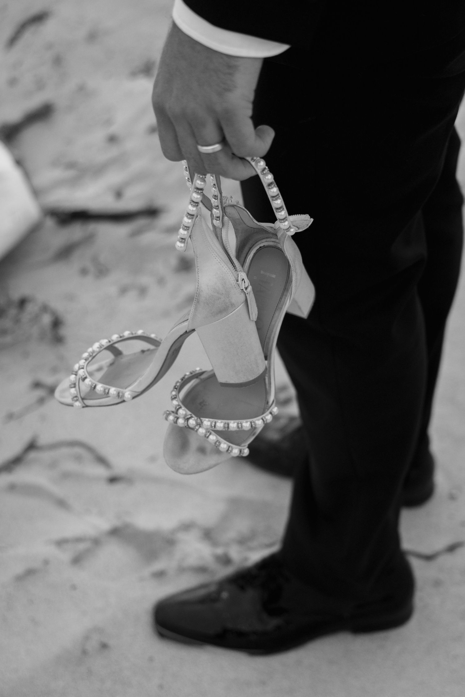 A hand holds elegant pearl-studded bridal heels against a contrasting black suit pants leg in black and white.