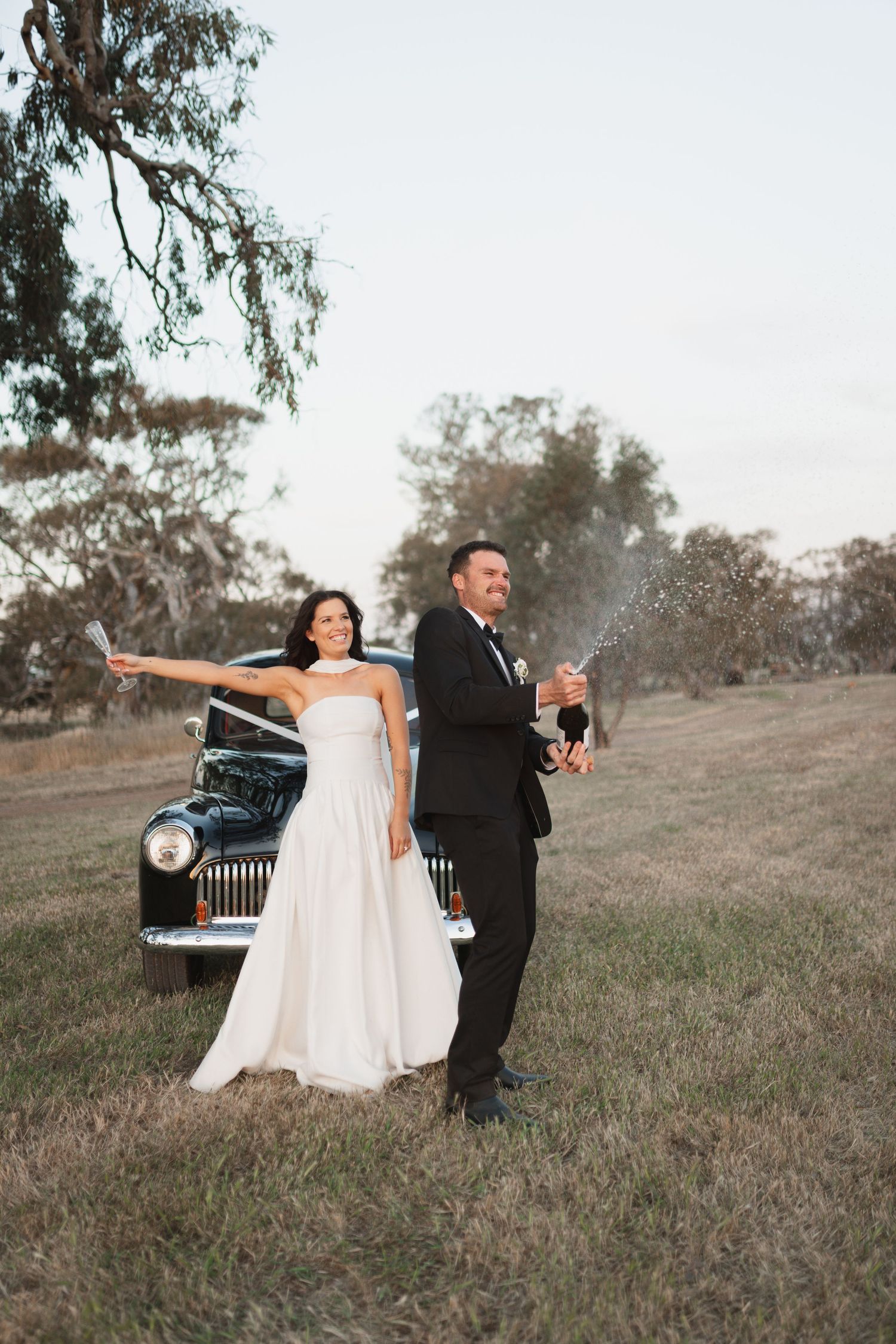 Bride and groom celebrating with champagne by a vintage black car in a rustic field setting.