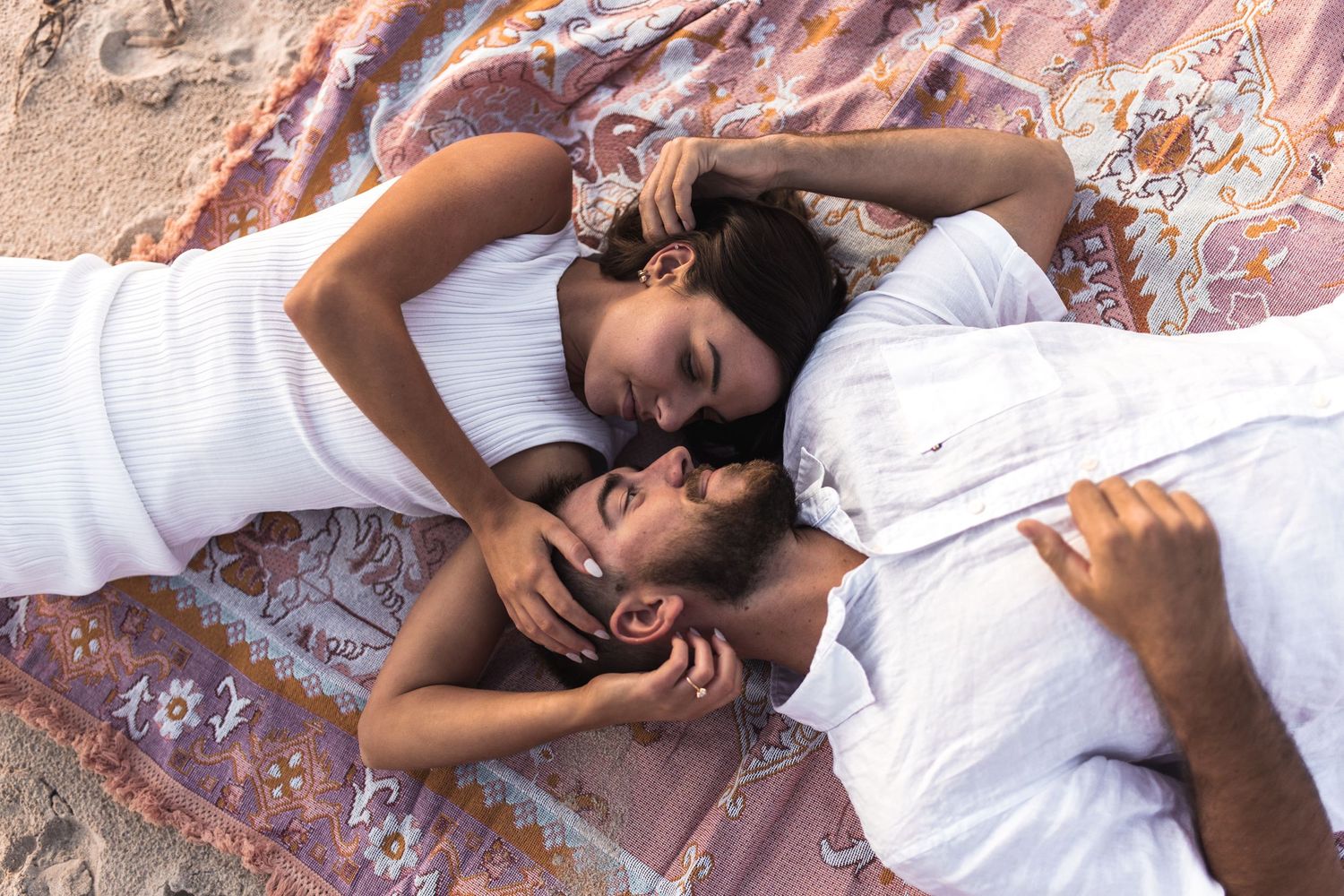 Couple lying on a patterned blanket at the beach wearing white outfits.