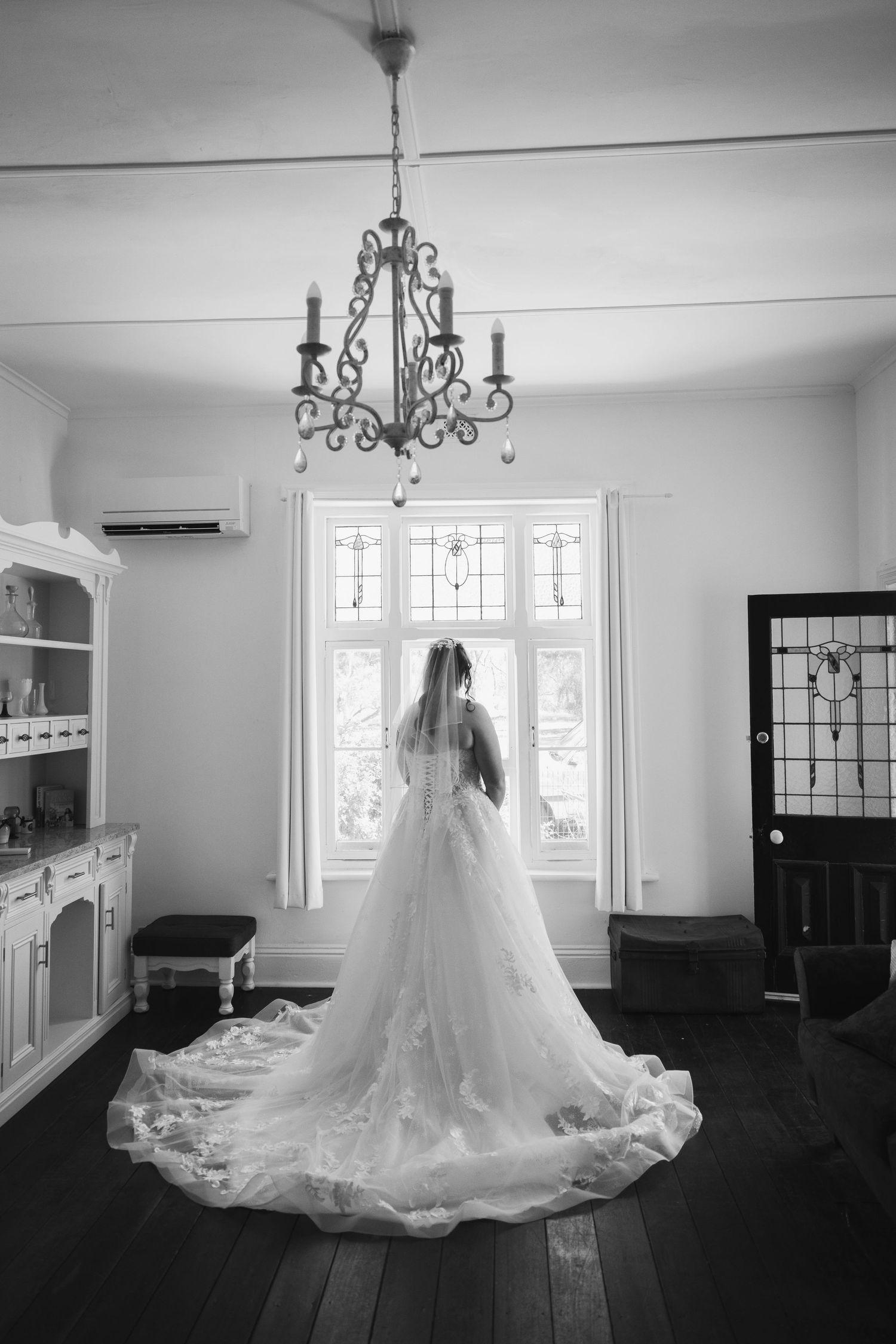 A bride in a flowing white wedding gown with long train stands silhouetted against bright windows under an ornate chandelier.