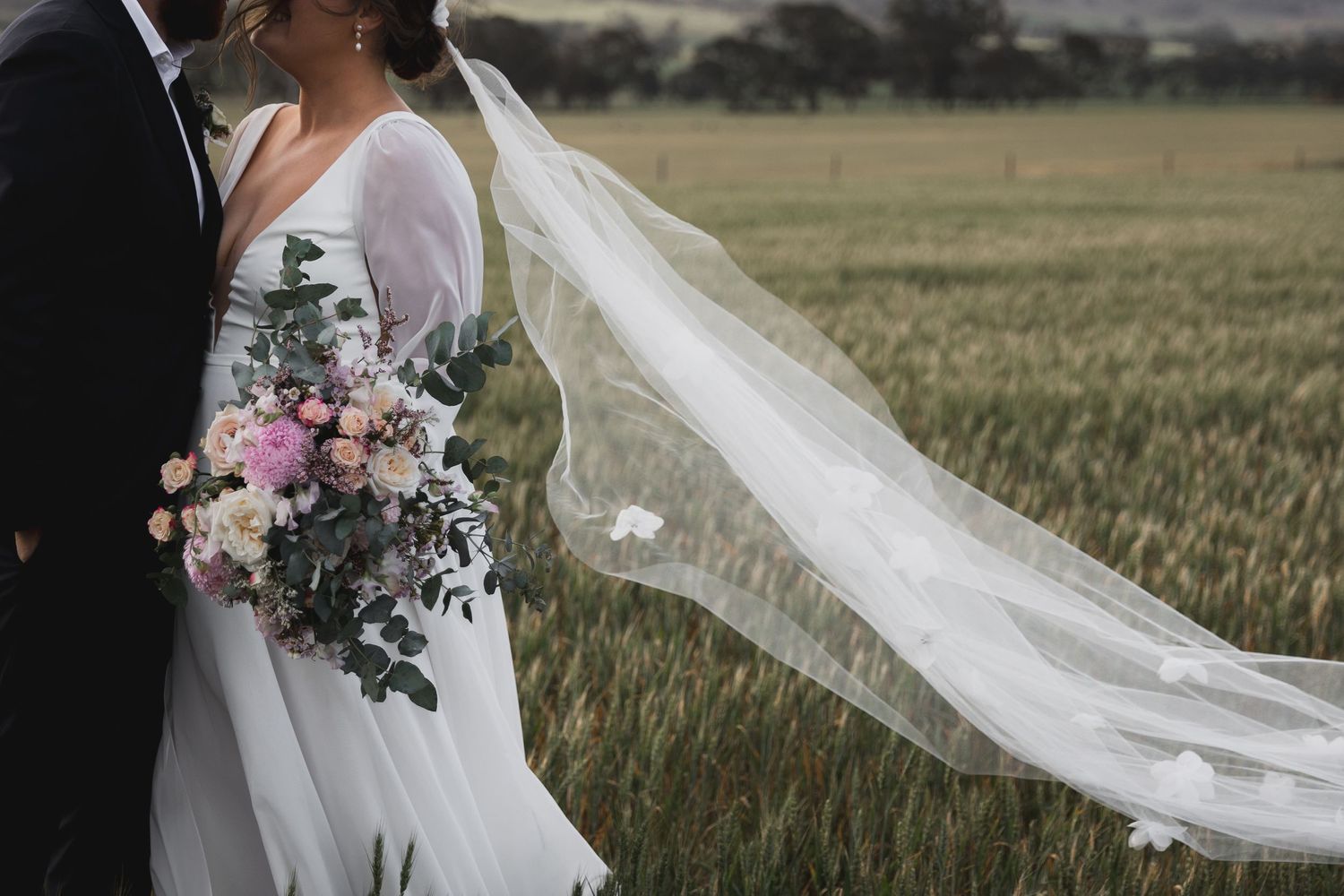 Elegant wedding couple with flowing veil and pink bouquet in a grassy field.
