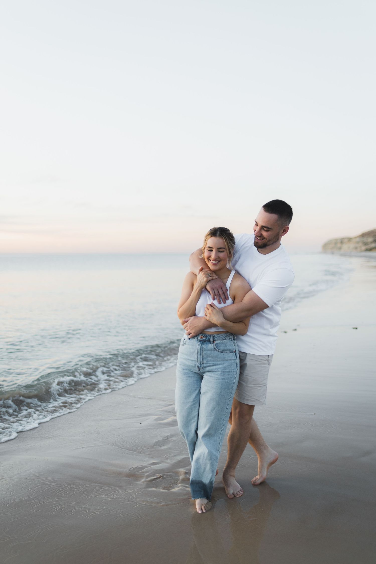 Couple embracing on beach at sunset wearing casual white and denim outfits.