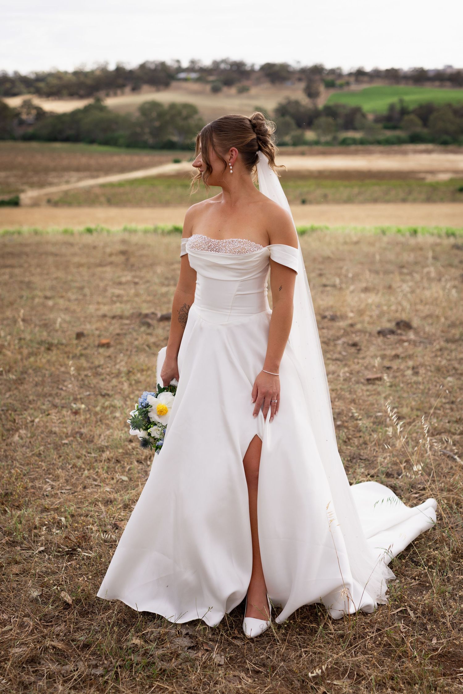 Bride in off-shoulder white gown with slit holds bouquet in rustic field, veil flowing, rolling hills behind.