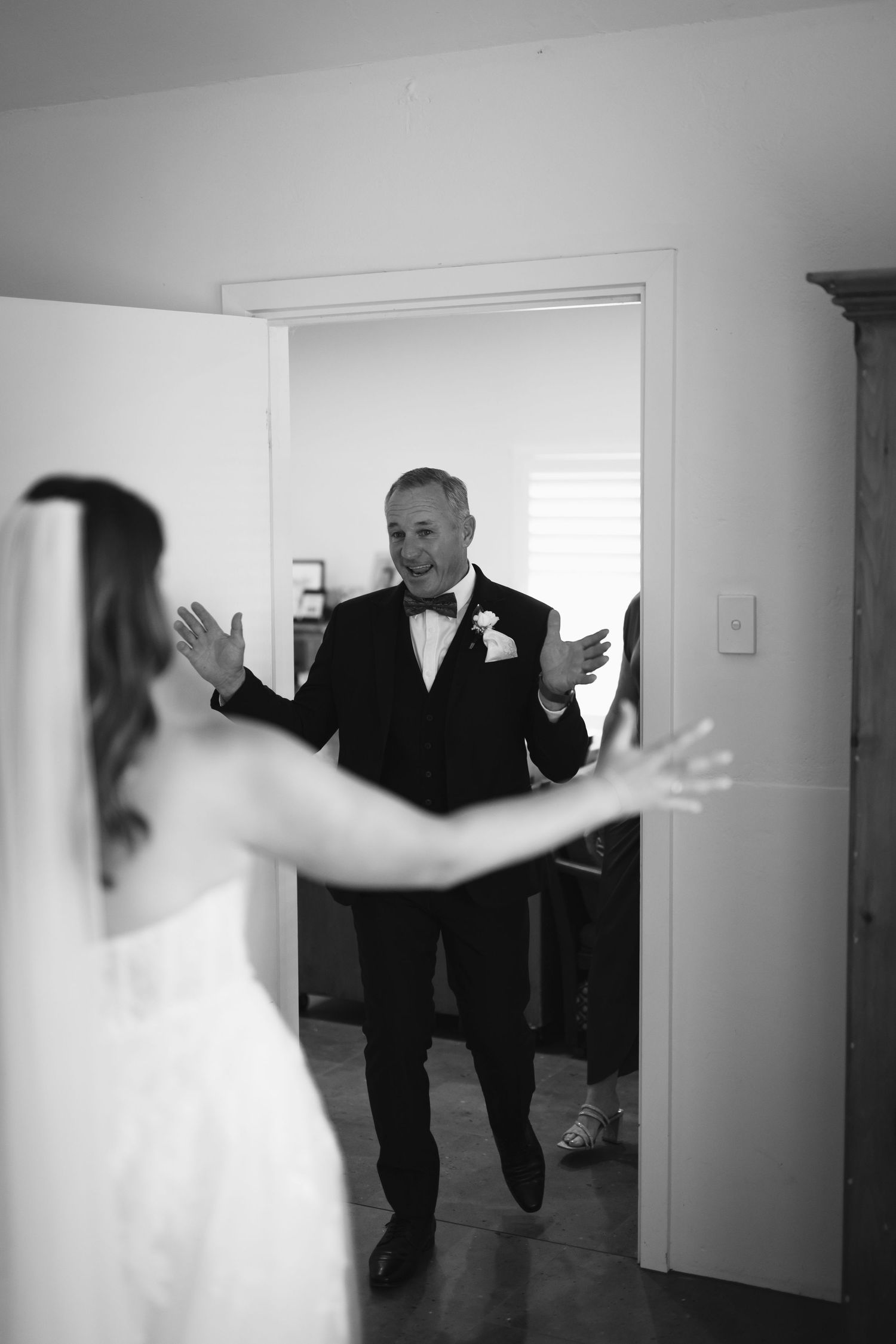 A heartwarming black and white wedding moment as someone in a tuxedo enters through a doorway, met with open arms.