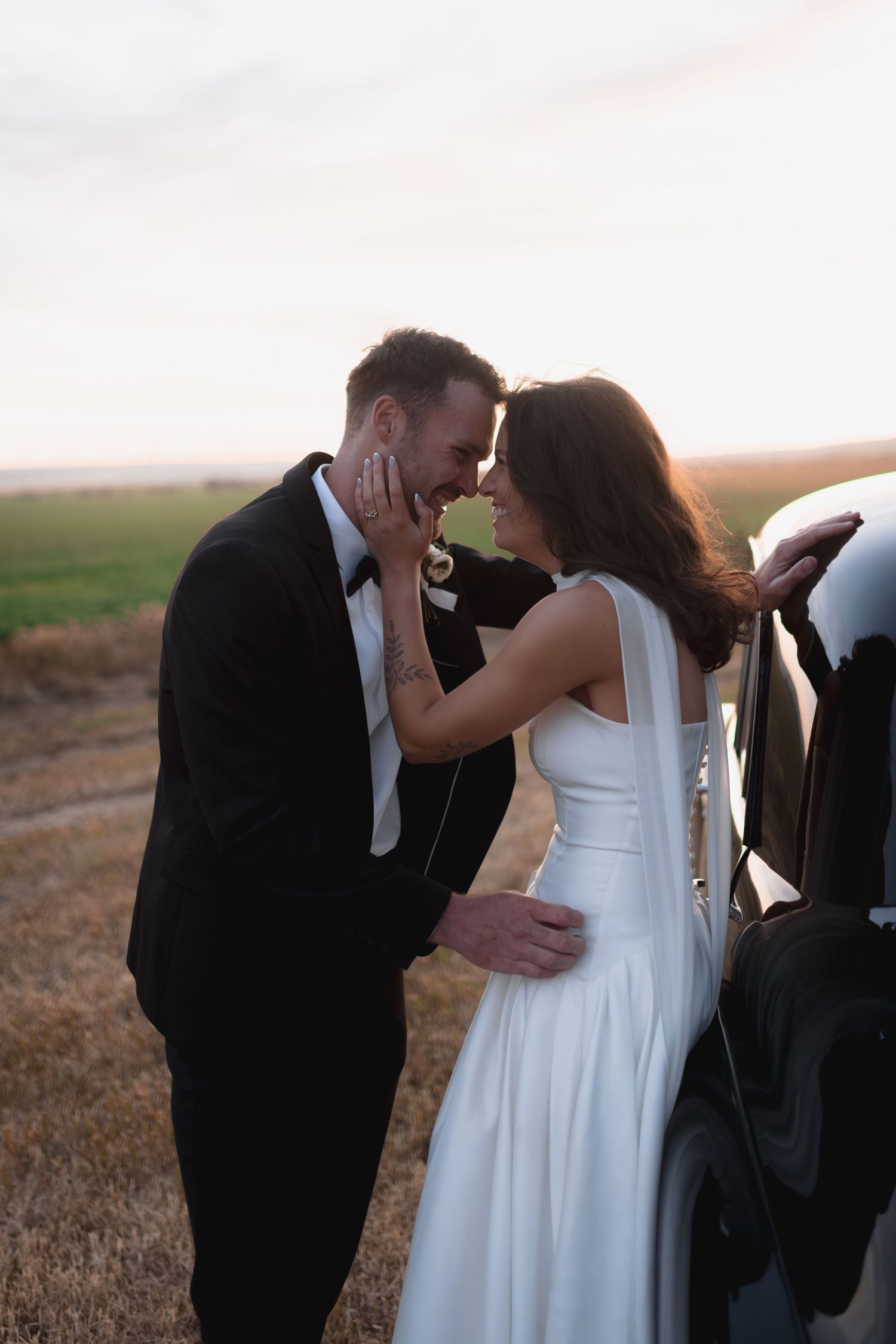 Bride and groom share romantic moment leaning against car in golden field at sunset, bride in white gown, groom in black tux.