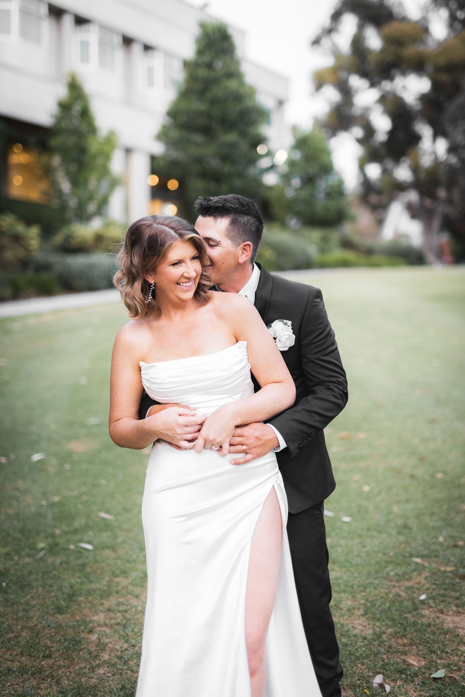 A joyful wedding couple shares a loving embrace and laughter on a grassy lawn with trees in the background.