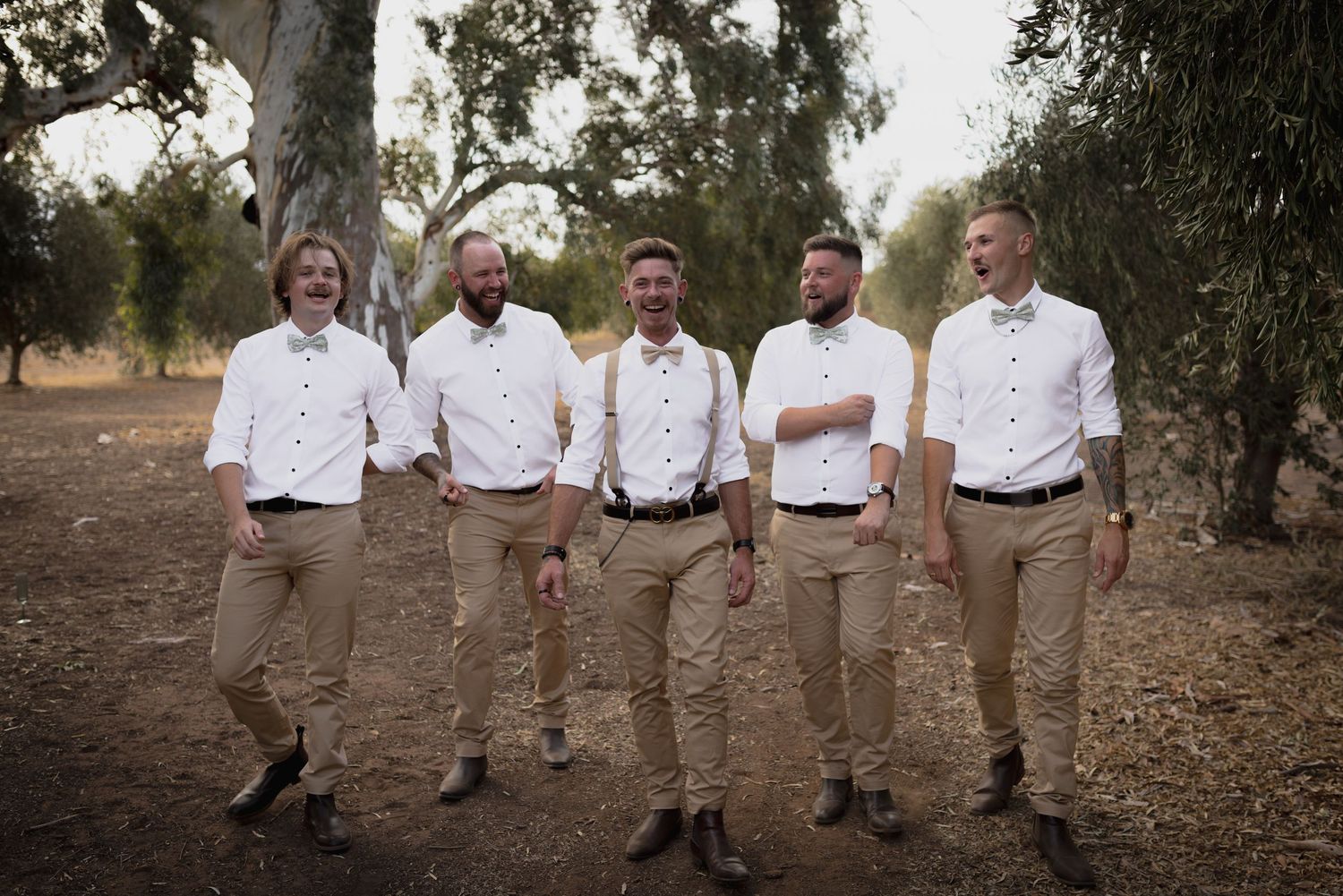 Wedding party wearing matching tan pants and white shirts pose together outdoors under eucalyptus trees.