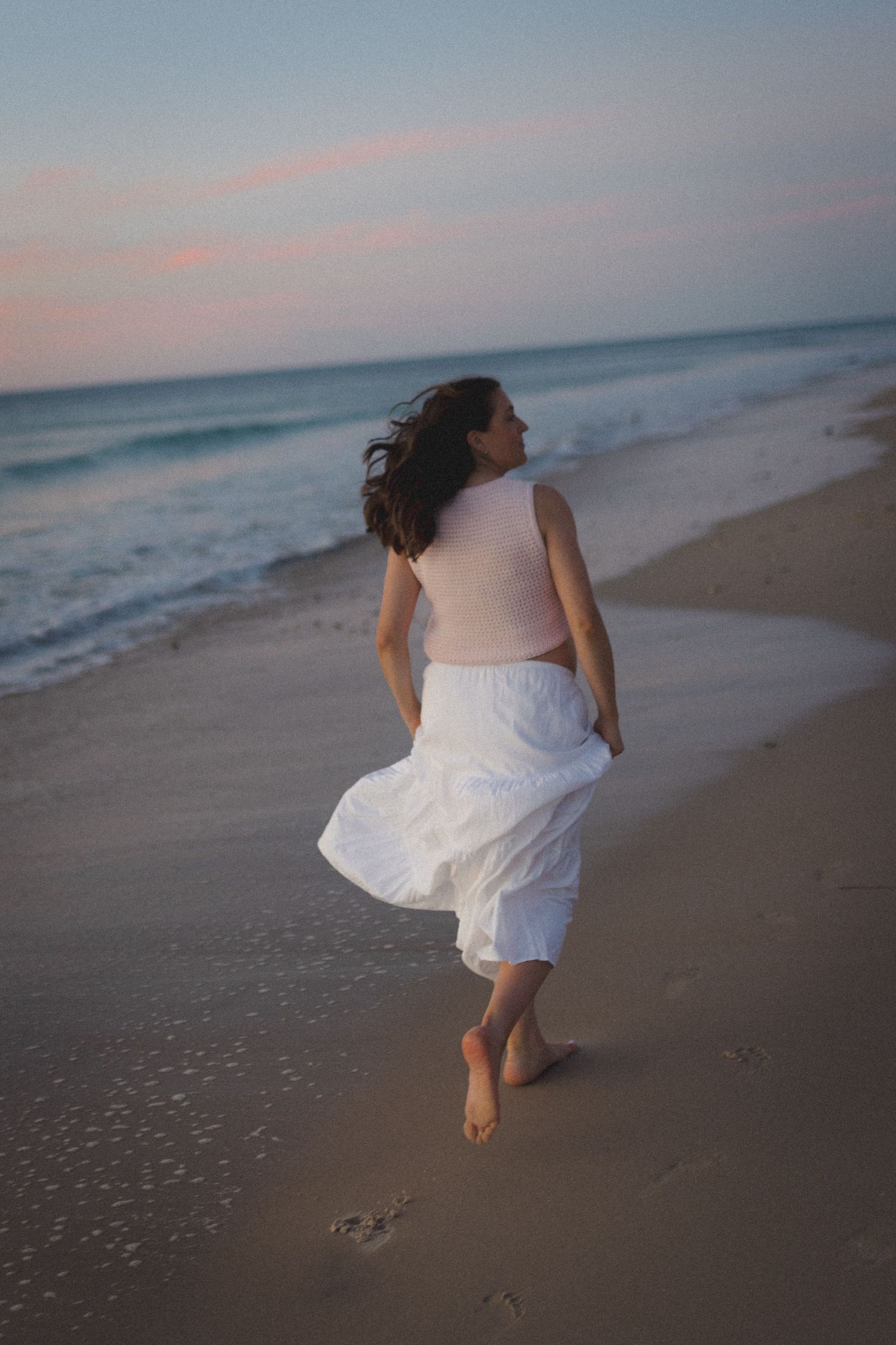 A barefoot figure in a white dress walks along a beach at sunset with waves crashing in the background.