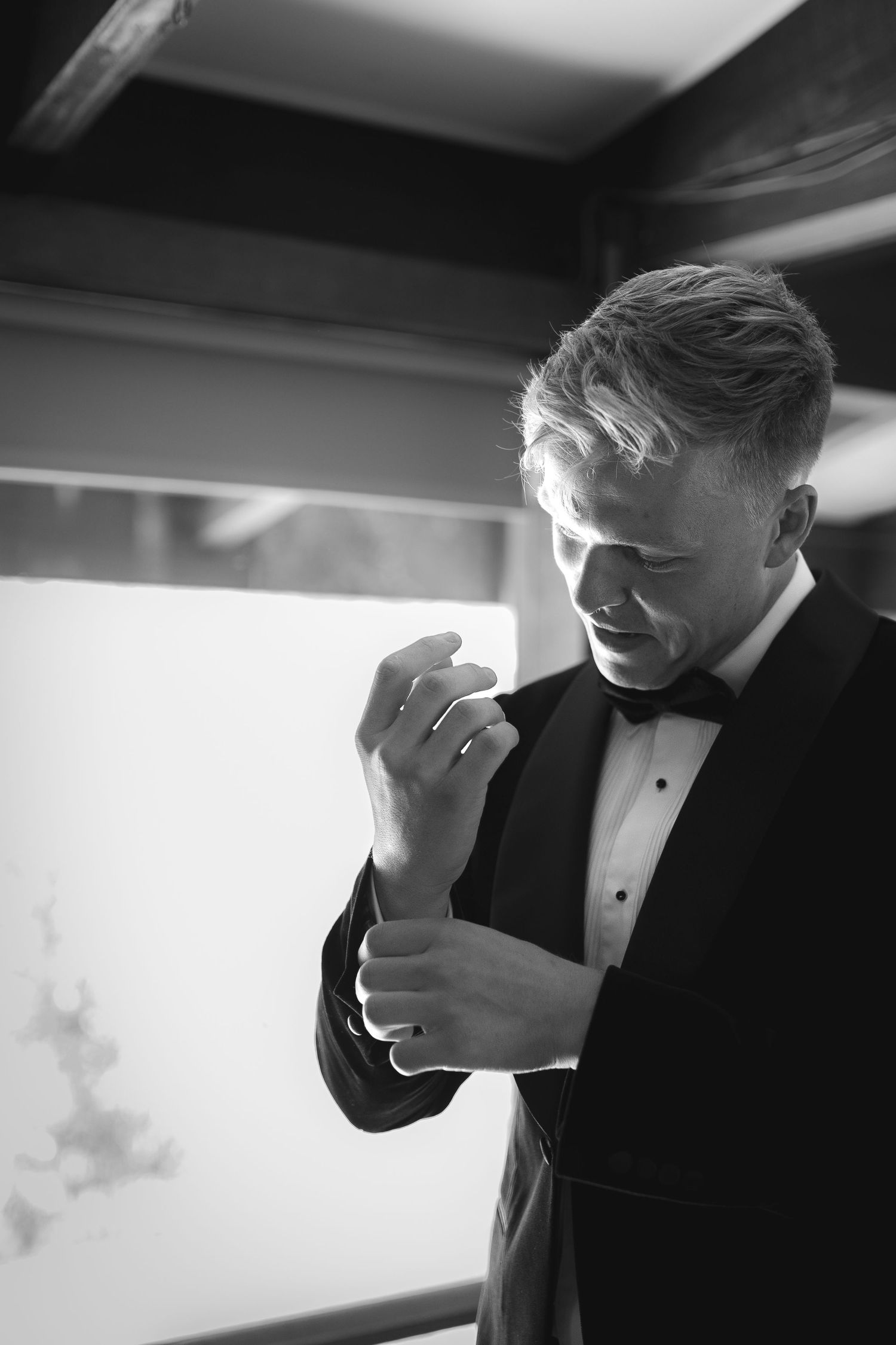 A groom in a black tuxedo adjusts his cuff in an elegant black and white portrait.