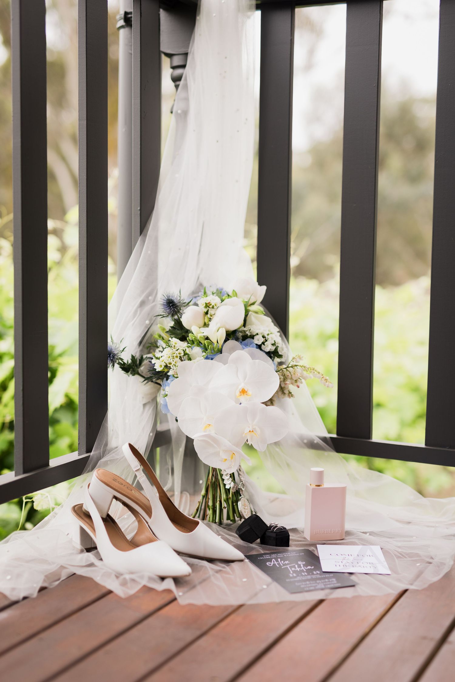 Bridal flat lay with white orchid bouquet, slingback heels, veil, pink perfume, and wedding stationery on a wooden deck.