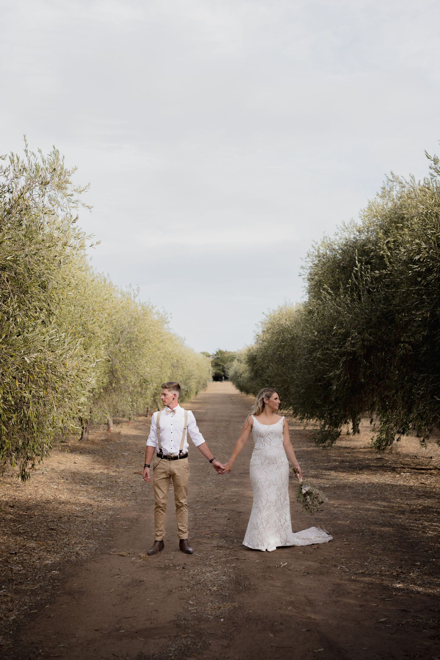 A couple holding hands while walking down a dirt path between rows of olive trees on their wedding day.
