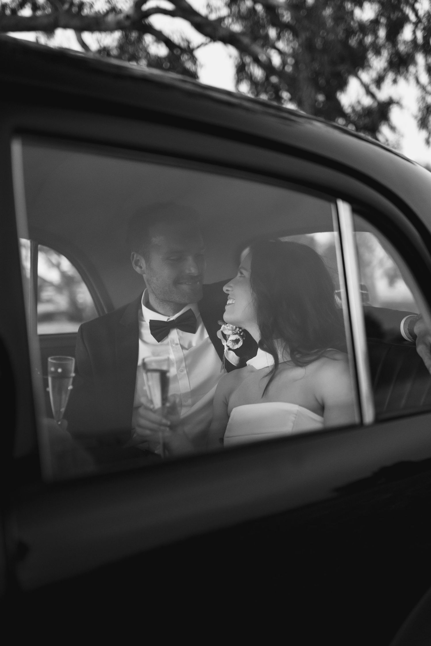 Black and white photo of a bride and groom sharing a romantic moment in a car, holding champagne glasses.