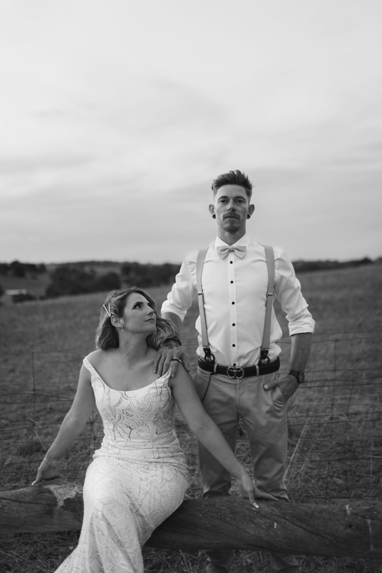 Black and white wedding portrait of couple in rustic outdoor field setting with bride seated and groom standing.
