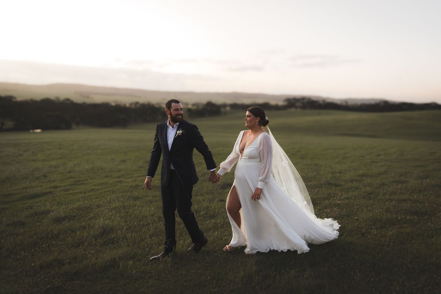Couple walking hand in hand through a green field at sunset in wedding attire.