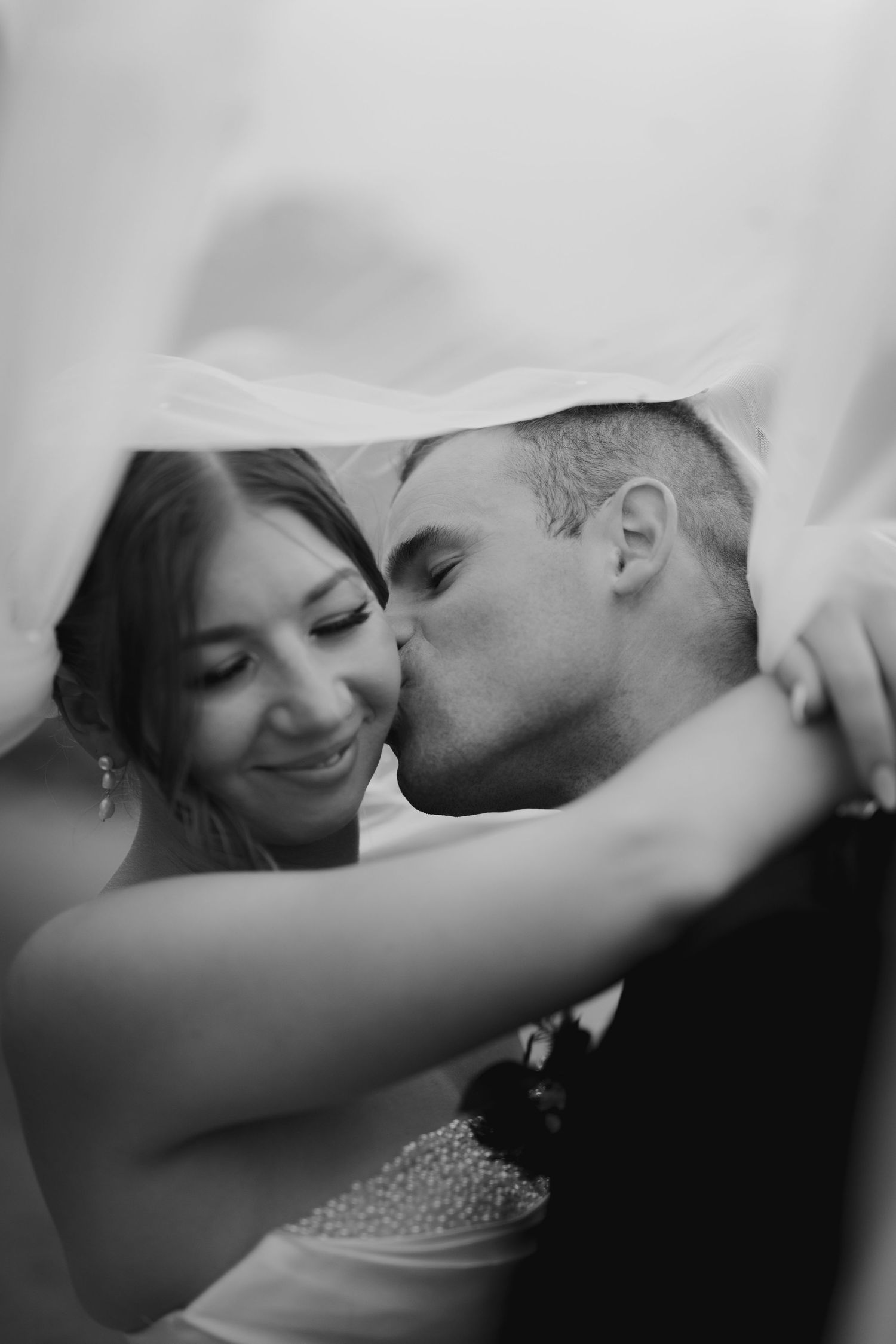 Black and white wedding photo of groom kissing bride on cheek under veil, bride smiling romantically.