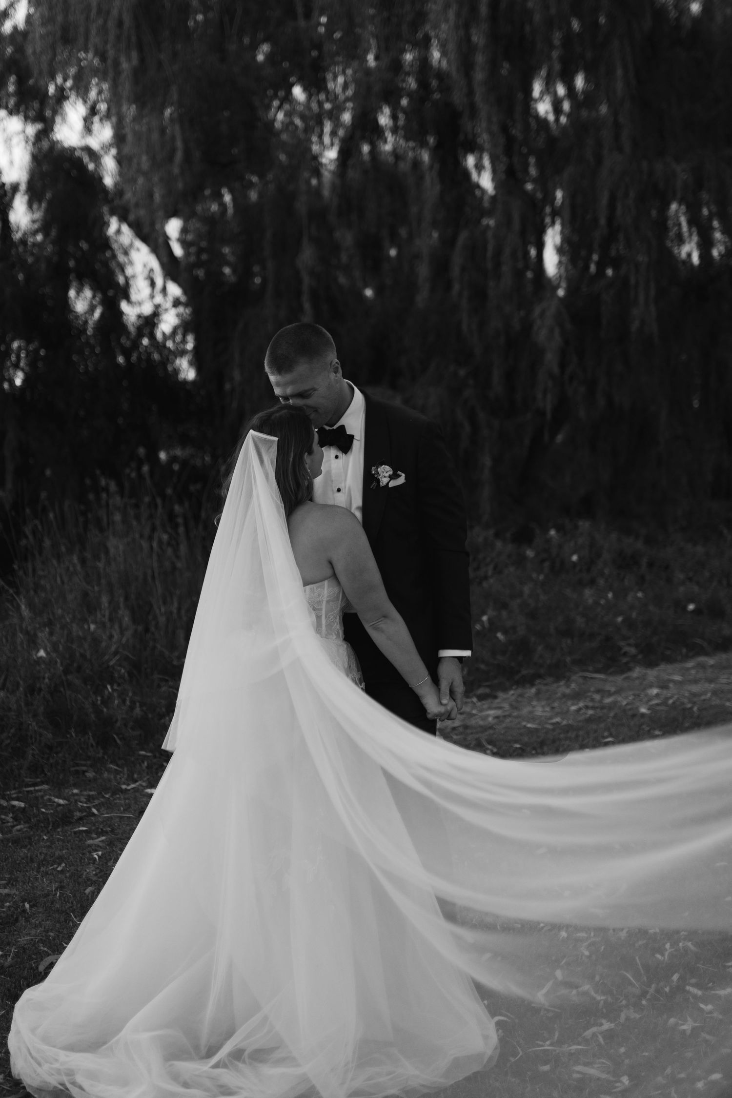 A romantic black and white wedding photo showing a bride's flowing white dress train against a dark outdoor setting.