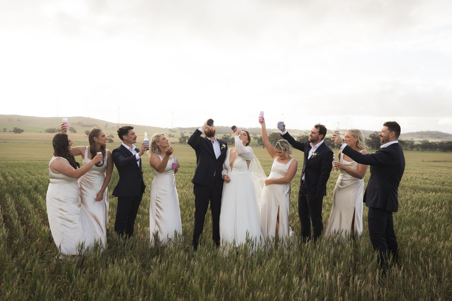 Wedding party celebrates together on a grassy field with raised hands during golden hour photography session.