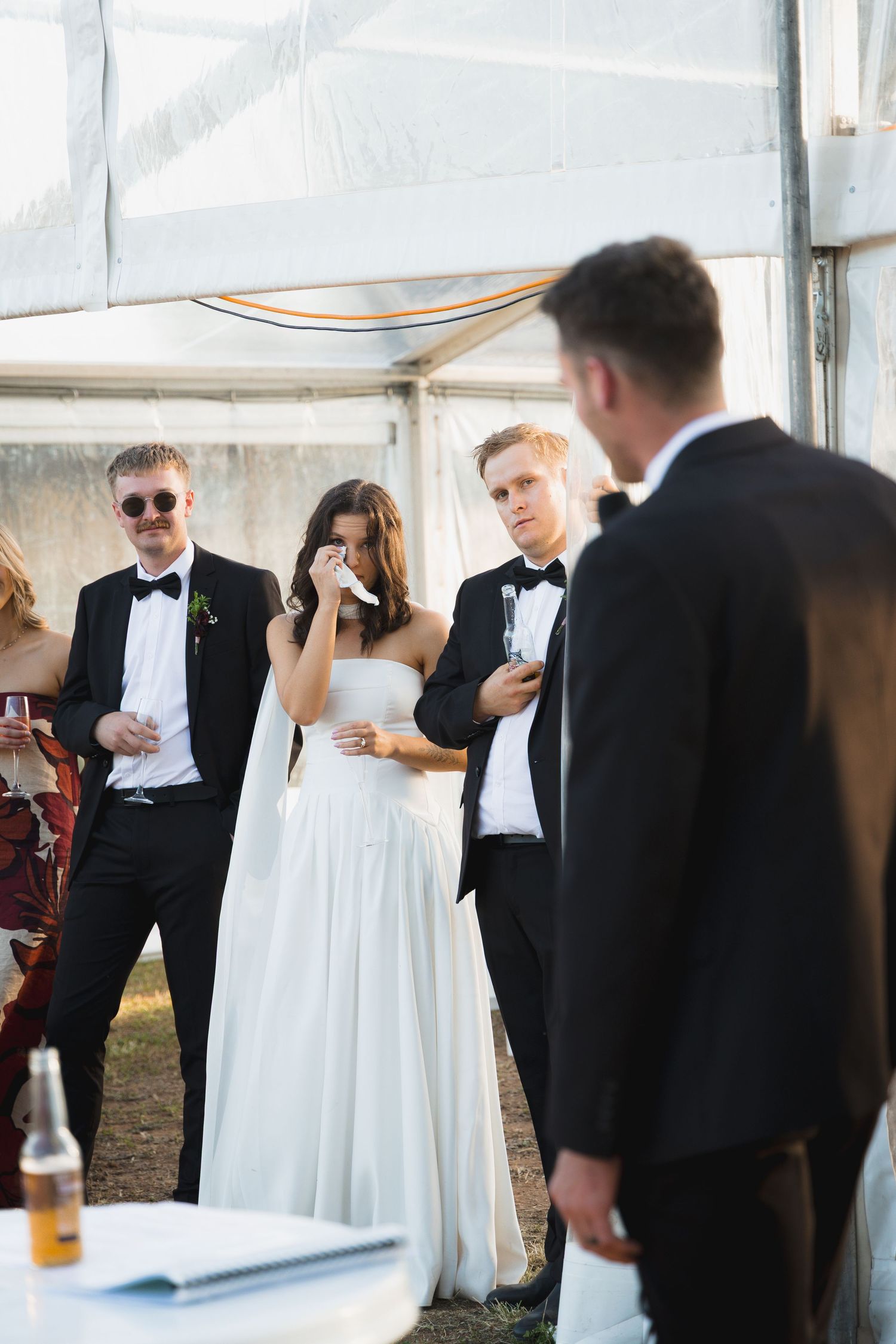 Bride wiping tears at wedding reception while groomsmen in tuxedos stand nearby under a tent.