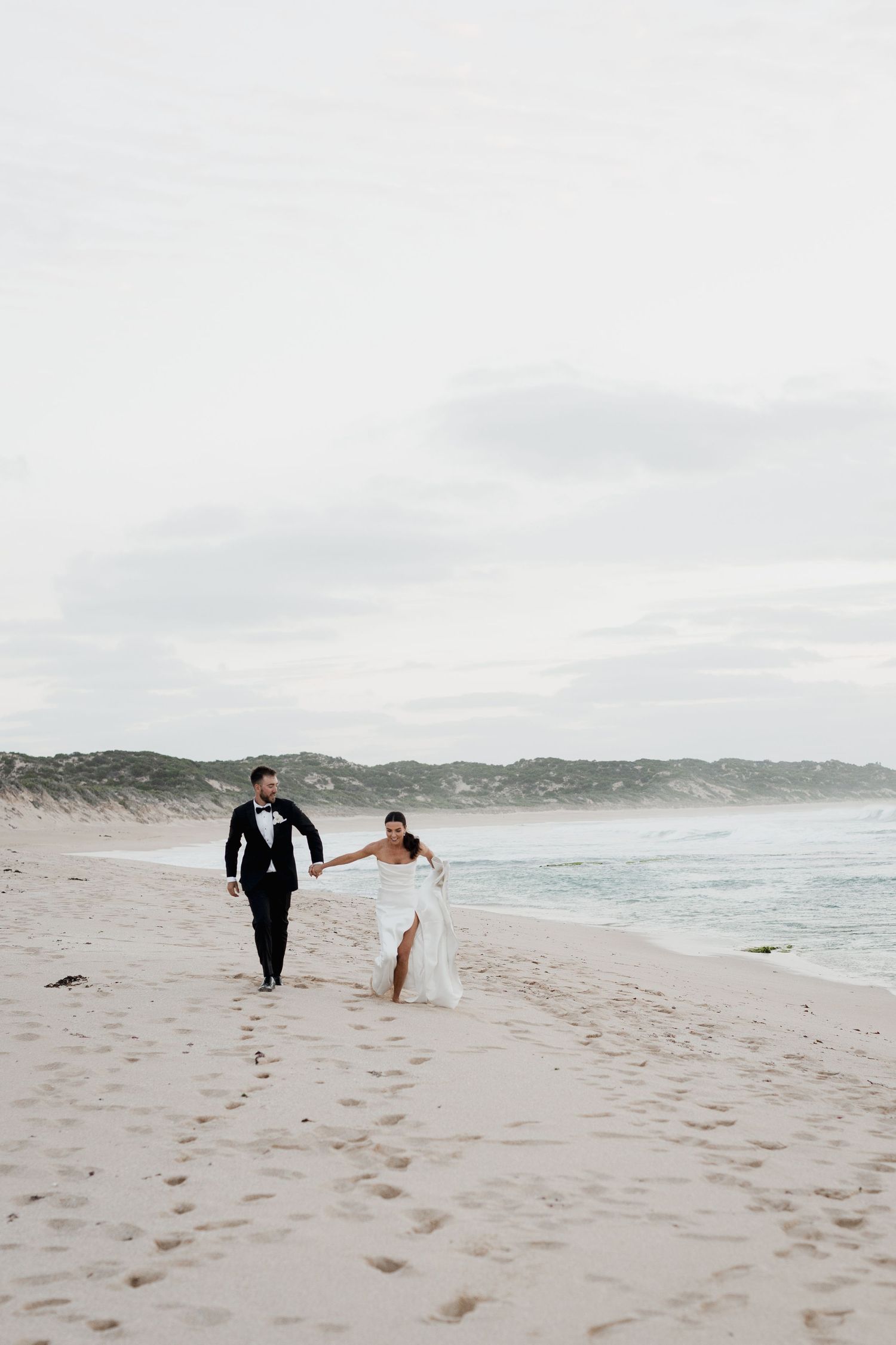 A couple walks hand in hand along a serene beach with rolling dunes and cloudy skies in the background.